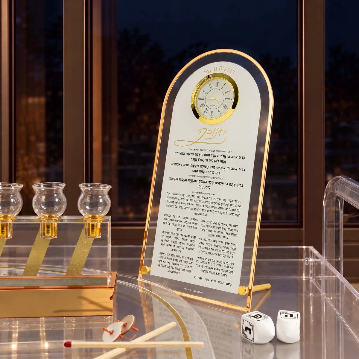 Chanukah brachos clock from Waterdale Collection, frosted Lucite in a gold arch frame with a working clock above the Chanukah brachos set up by night next to a window and menorah