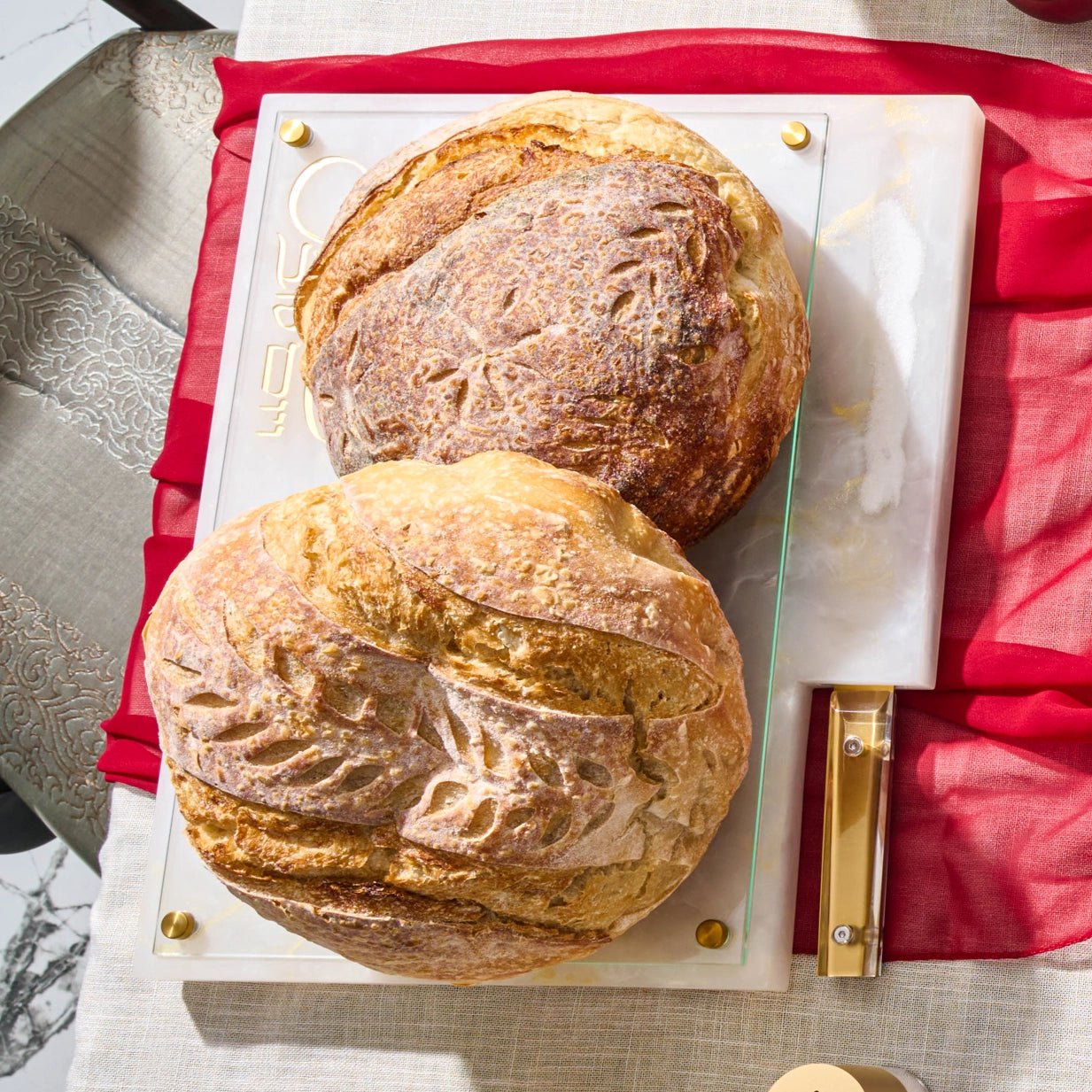 Two round artisan sourdough bread loaves displayed on aרקדןמ and glass serving platter with gold hardware, set on a red fabric runner over a light table.