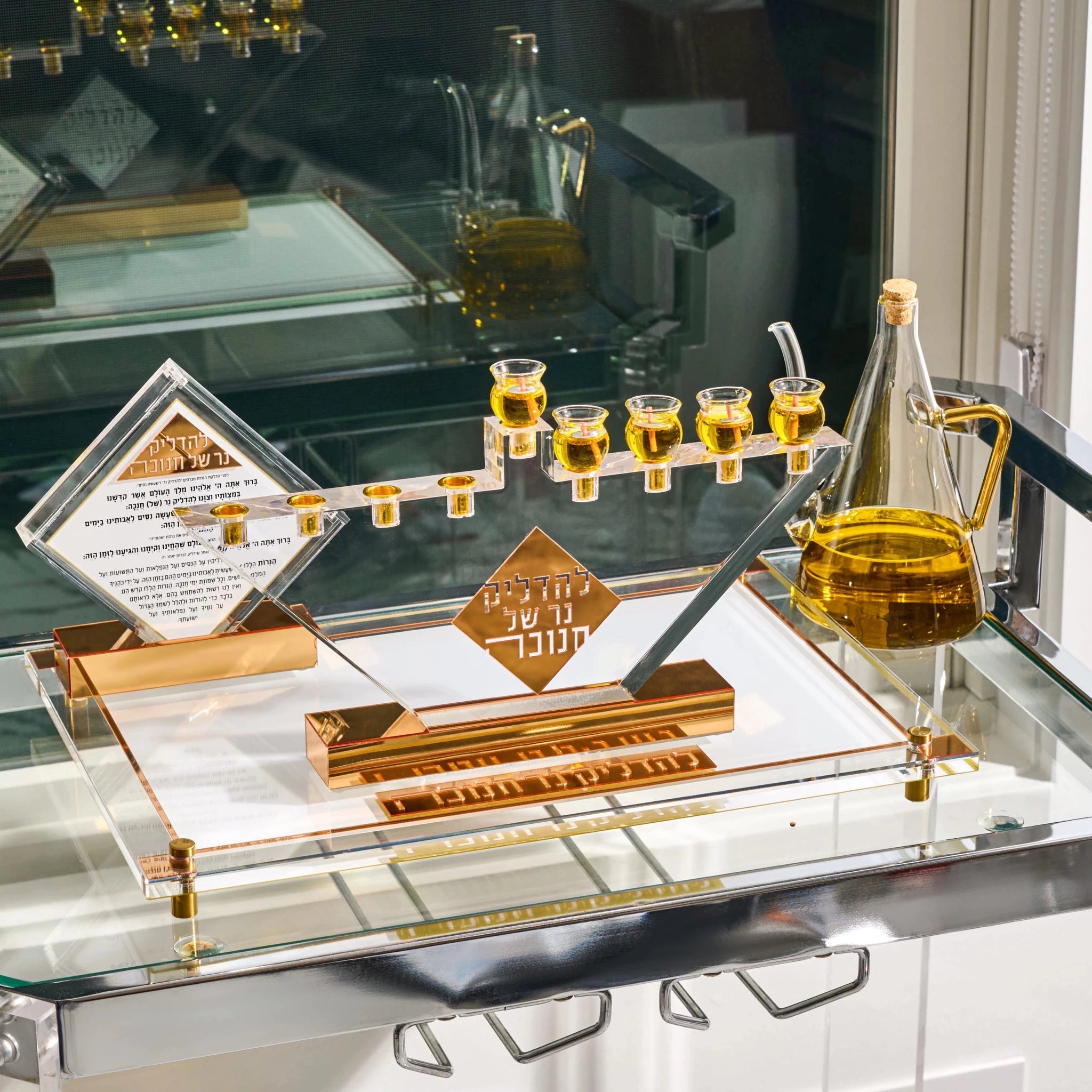 An acrylic and gold Hanukkah menorah set on a glass shelf, featuring a menorah with small clear cups filled with oil, a clear oil bottle, a diamond-shaped plaque with Hebrew and English text, and a central gold sign.