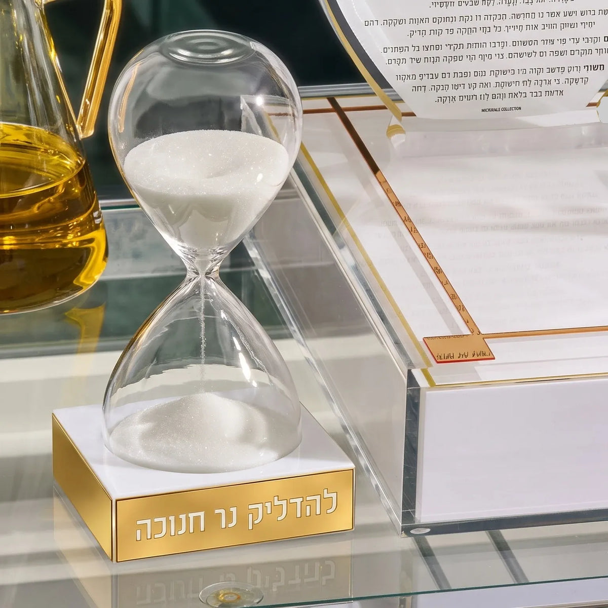 A clear glass hourglass for a chanuka timer with white sand, set on a polished gold rectangular base engraved with Hebrew text, set by a window for a chanuka display