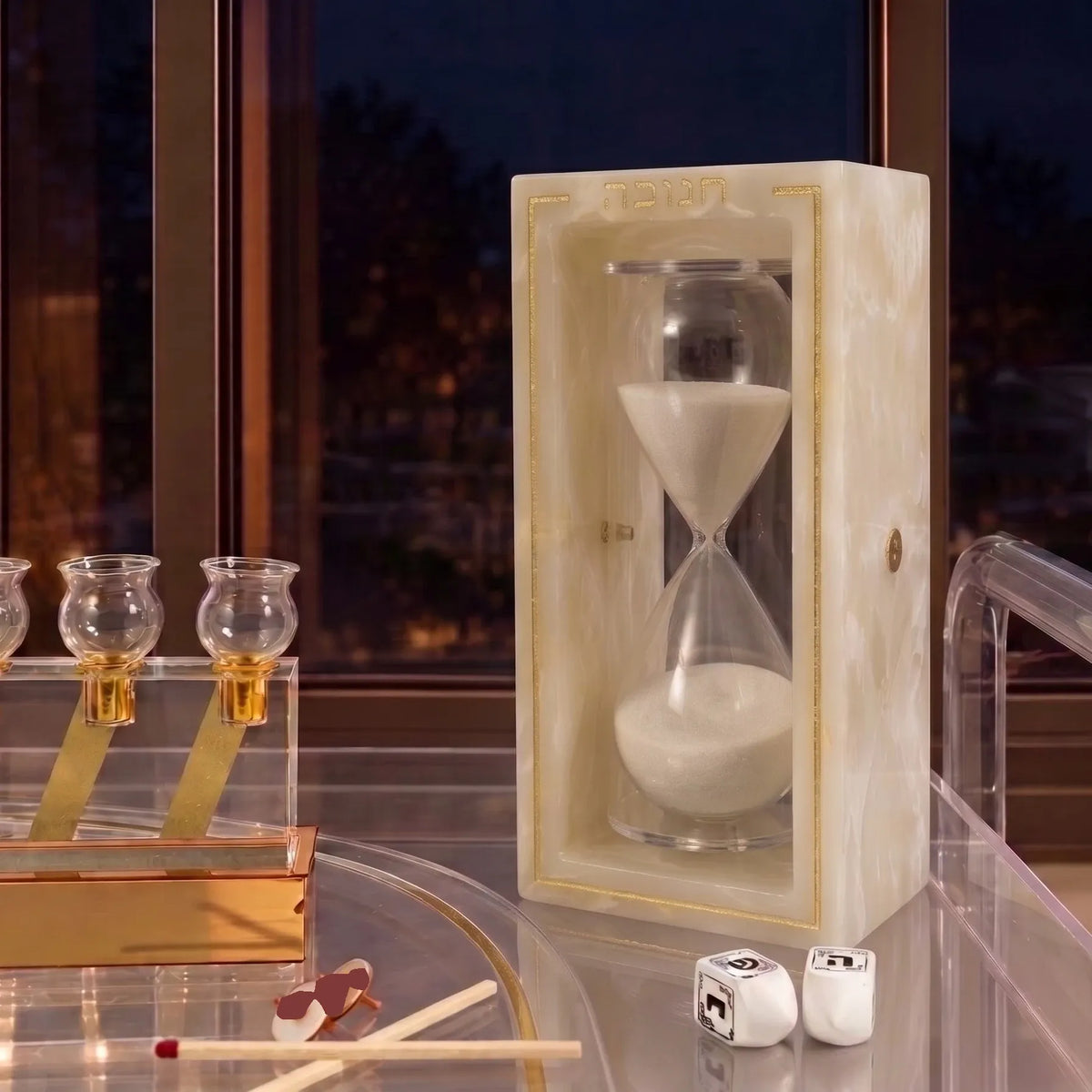 Gold resin Chanukah sand timer, a 30-minute glass hourglass in a resin frame lettered חנוכה for Chanuka set on a table near a window and a menorah