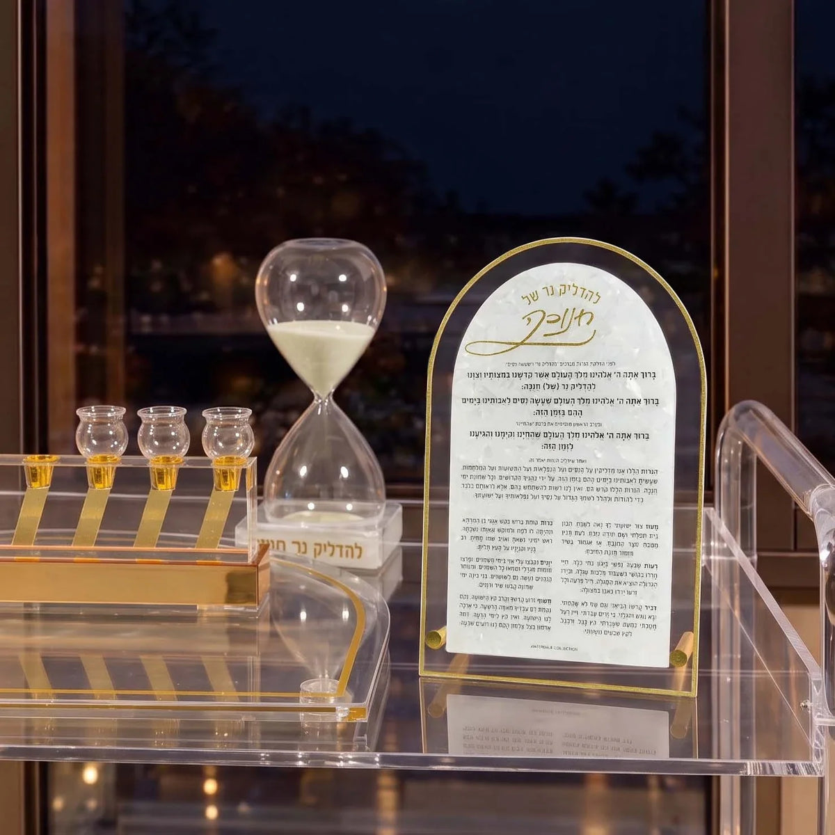 Pearl marble Chanukah brachos plaque beside a 30-minute sand timer and lit menorah on a clear Lucite cart at night