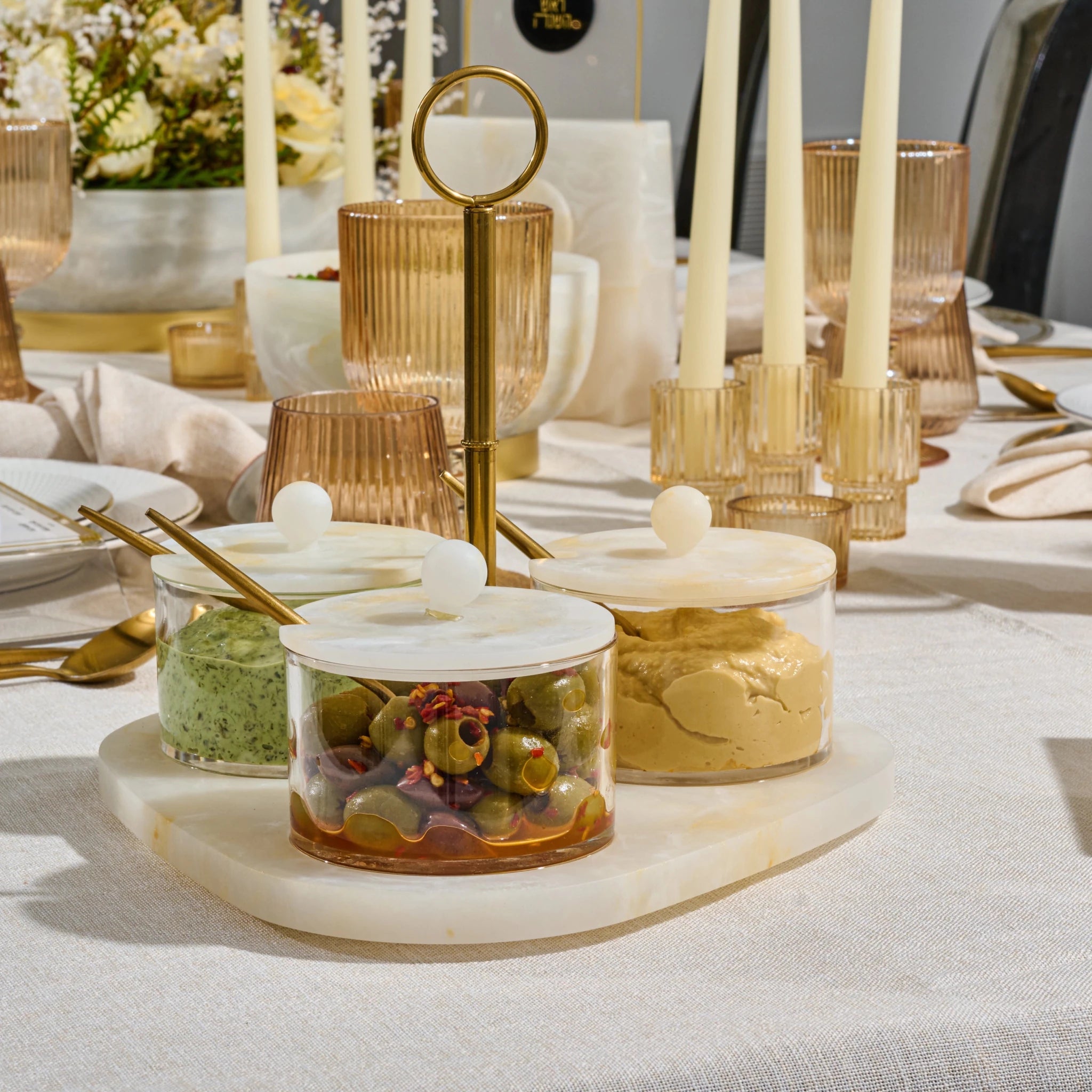 A styled tabletop setting with clear glass jars containing green olives and a beige spread, set on a resin tray with white lids and gold spoons. The scene includes amber glass candle holders with cream taper candles, ribbed amber glasses, and a white tablecloth in a bright, elegant dining environment.