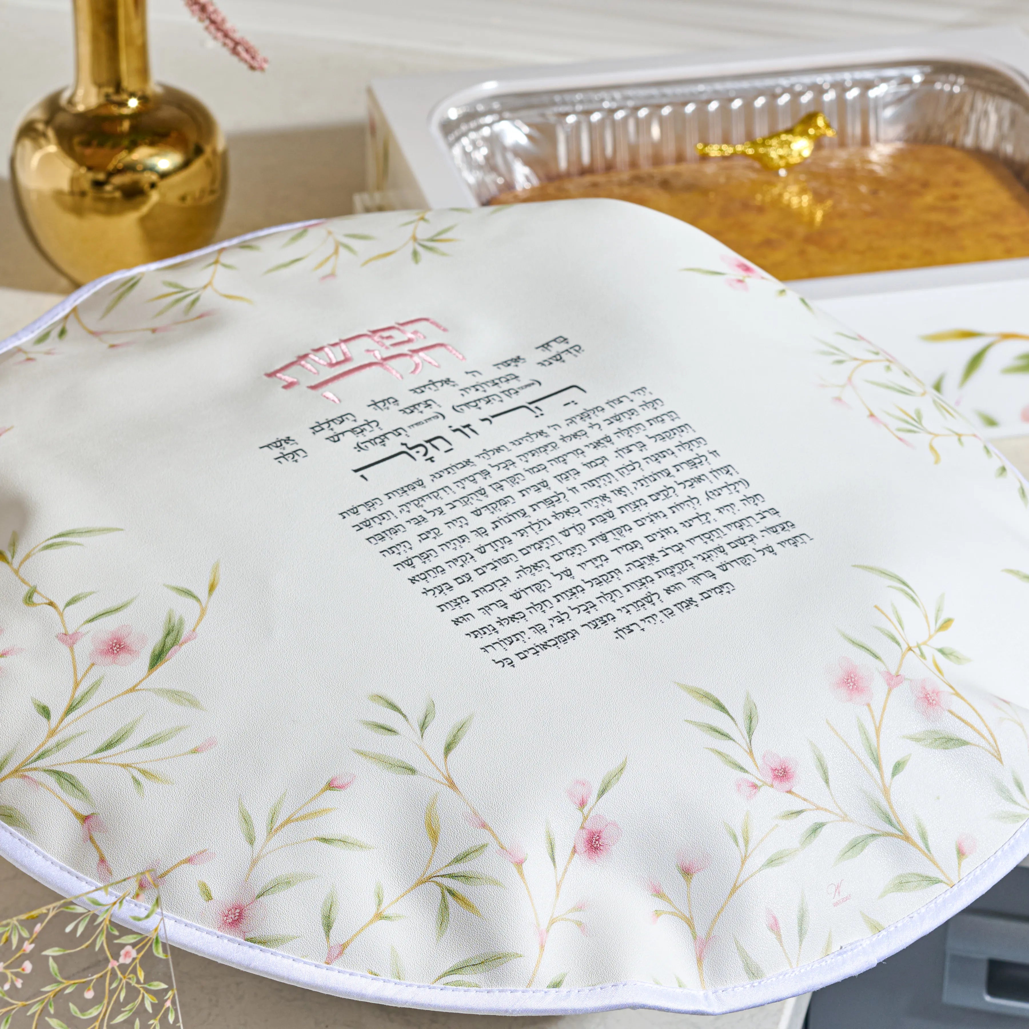 A round white Floral Chsllsh cover with printed Hebrew text and pink floral illustrations, shown on a table next to a gold-colored decorative bottle and a foil pan of leavened bread.