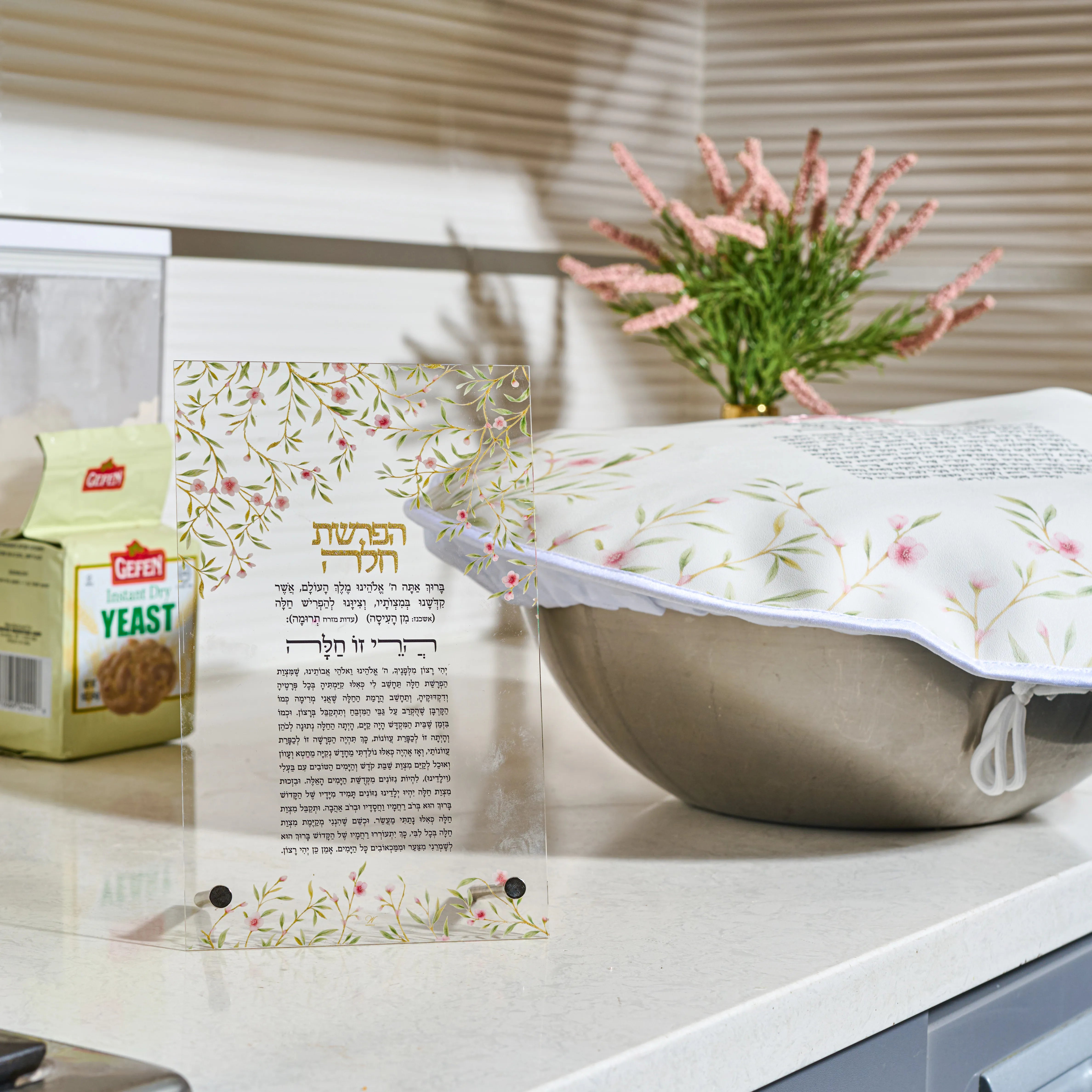 A decorative Challah dough cover over with Hebrew text and a floral border stands on a kitchen counter next to a box of Leavened yeast. In the background, a white electric blanket with a matching floral pattern is visible, along with a vase of pink flowers and window blinds.