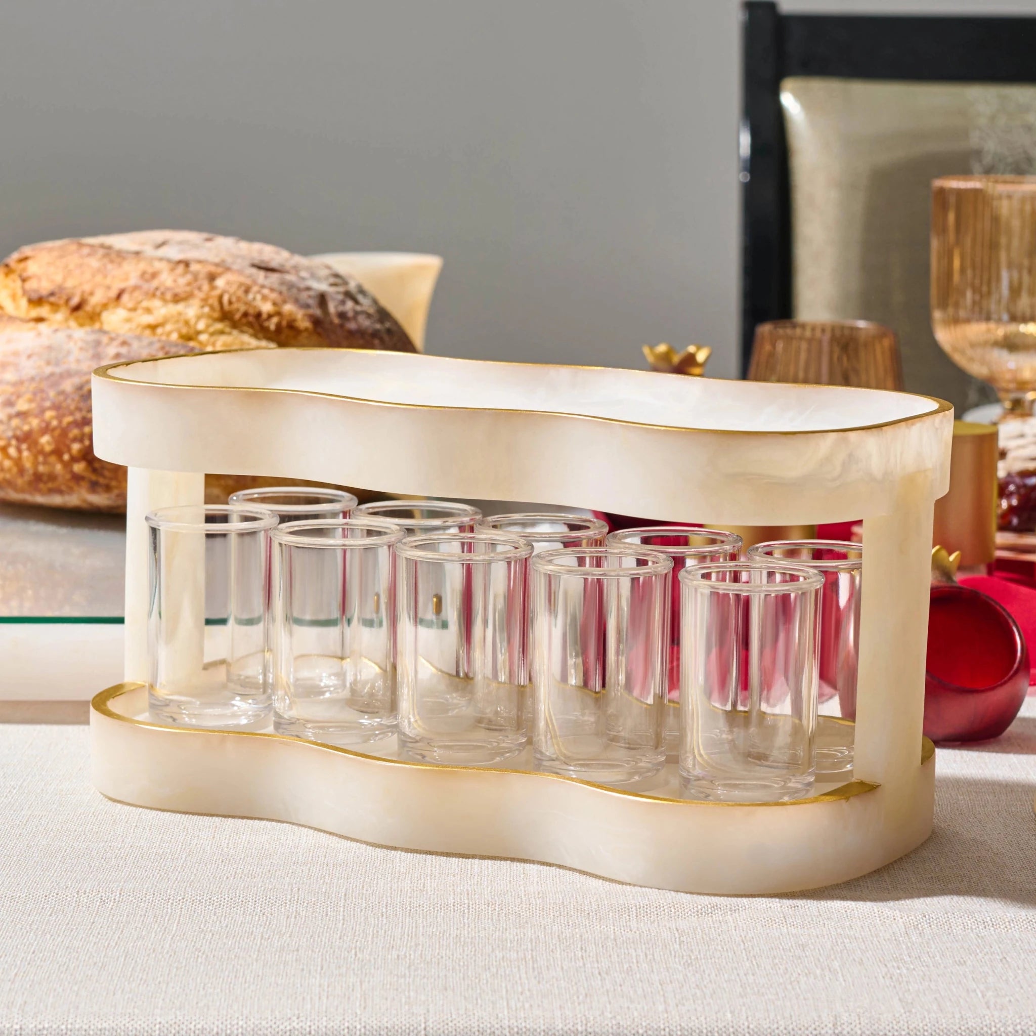A cream-resin shot glass holder with gold trim, holding multiple small clear shot glasses, styled on a table with bread and holiday decor in the background.