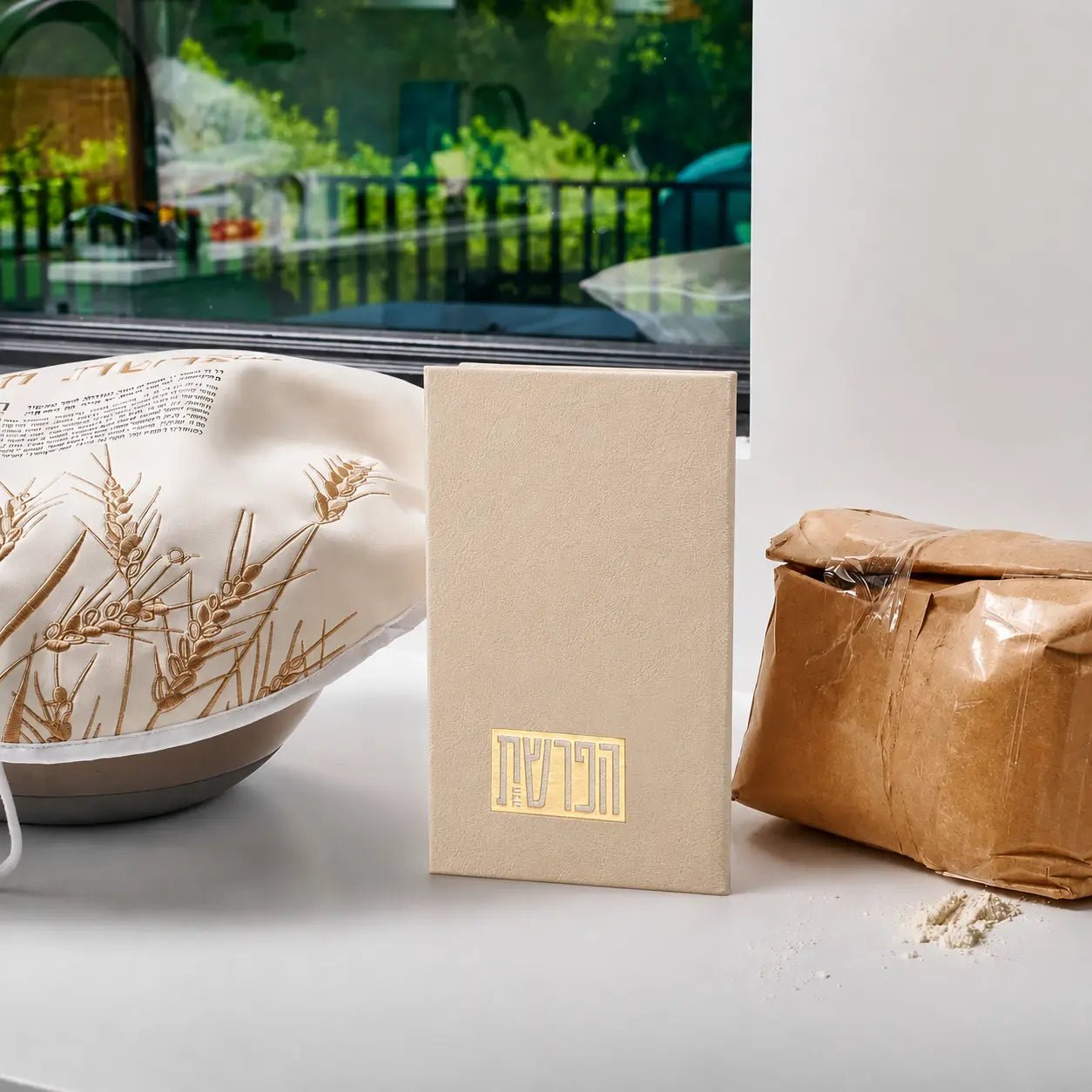 Tan leather Hafrashas Challah booklet displayed with dried wheat and kraft packaging on white surface near window.