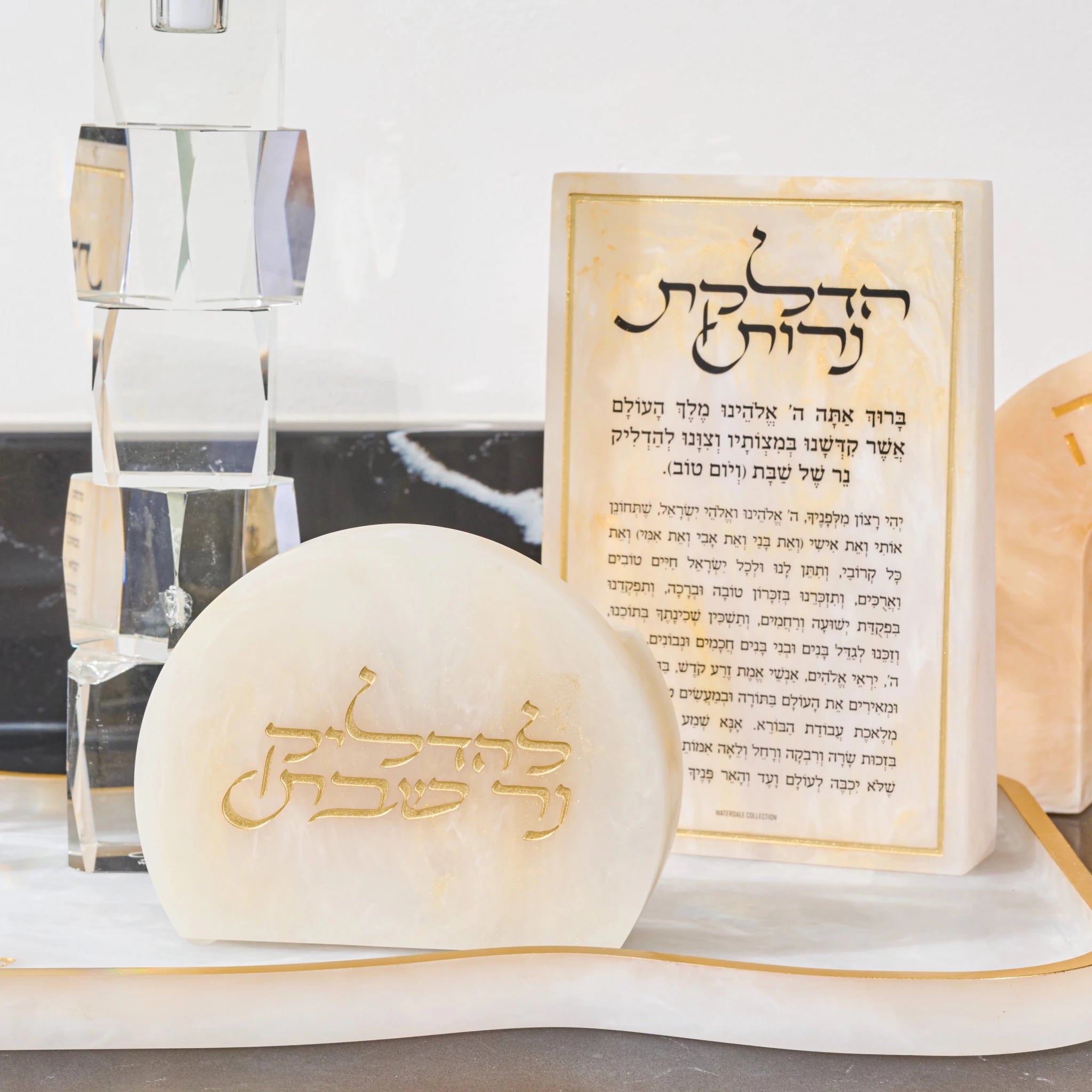 A small cream-colored resin plaque with gold Hebrew lettering sits in front of a matching resin matchbo displayed on a white marble surface