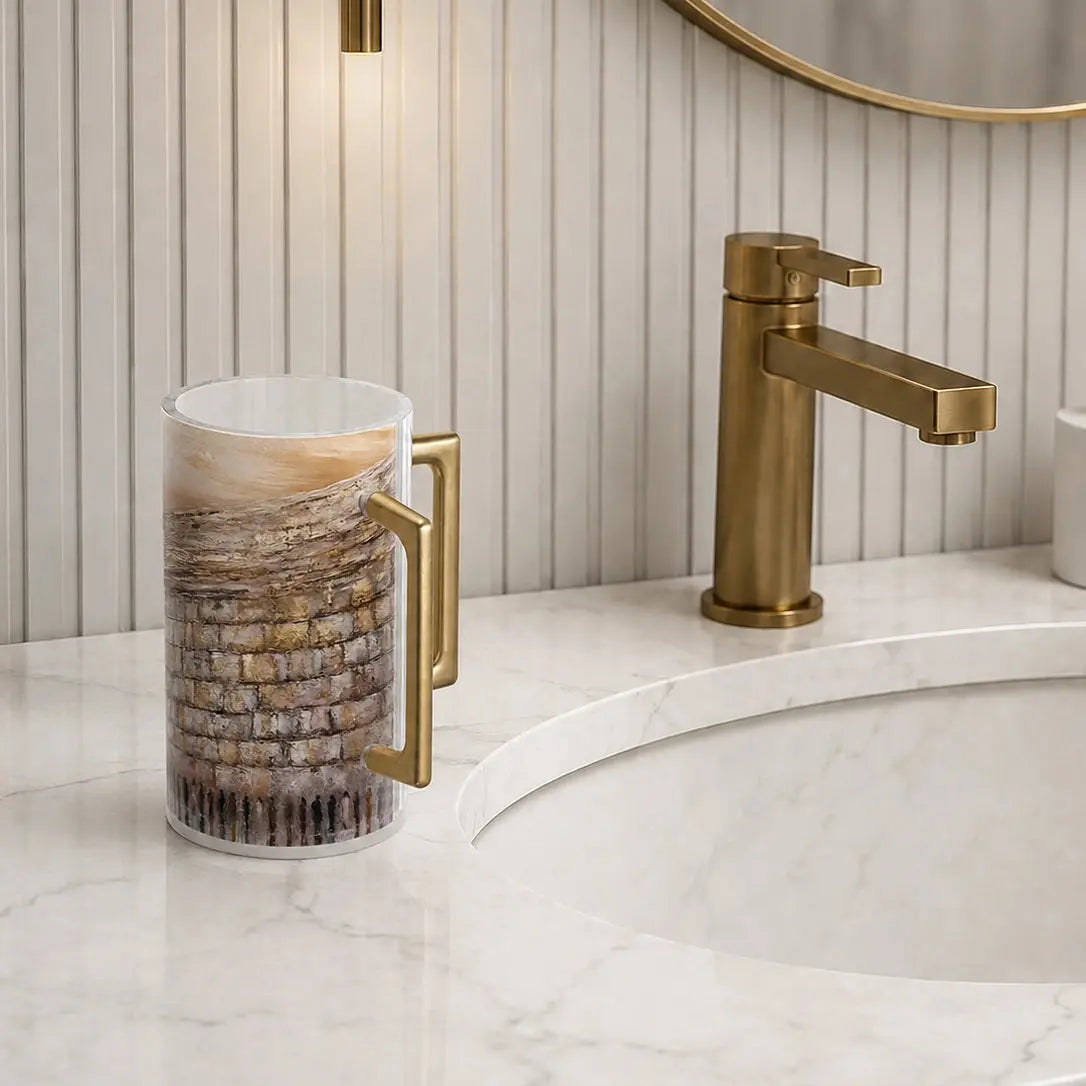 Clear Lucite Netilat Yadayim washing cup by Waterdale Collection, painted in taupe and gold Kotel artwork with a brushed gold handle, on a marble vanity.