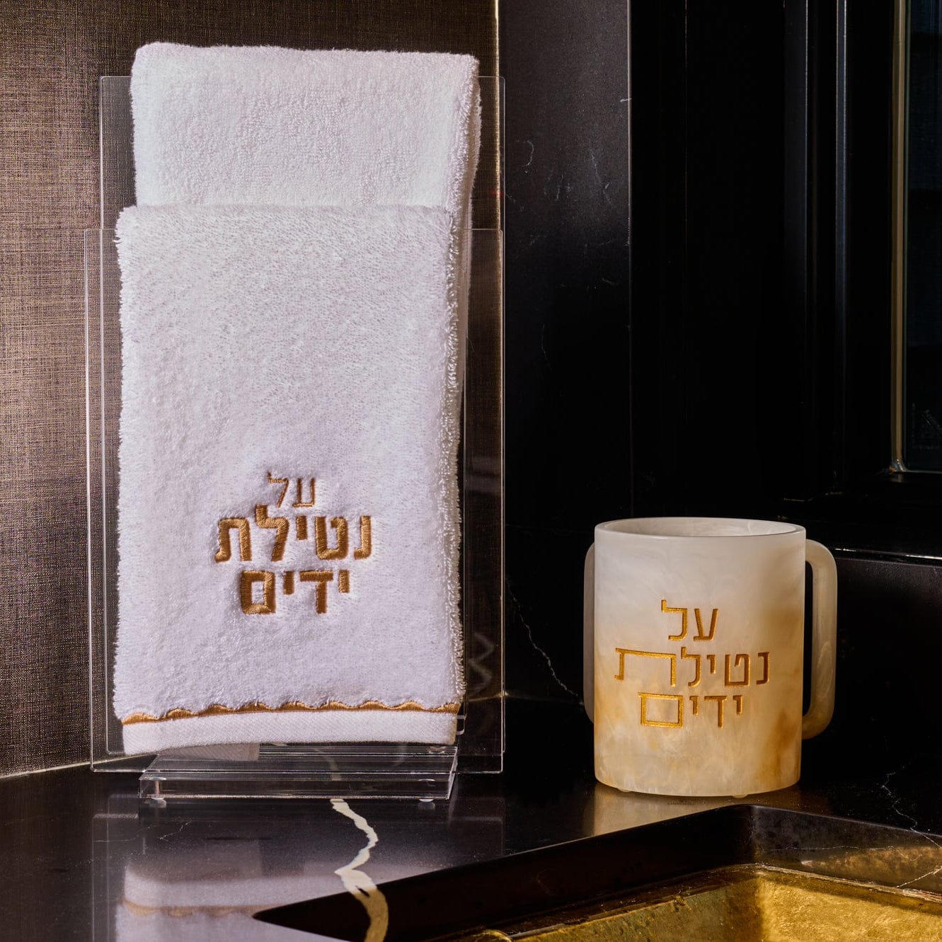 Ombre resin washing cup with gold “Al Netilas Yadayim” Hebrew lettering, displayed beside a white embroidered hand towel on a sleek black marble countertop near a modern sink.