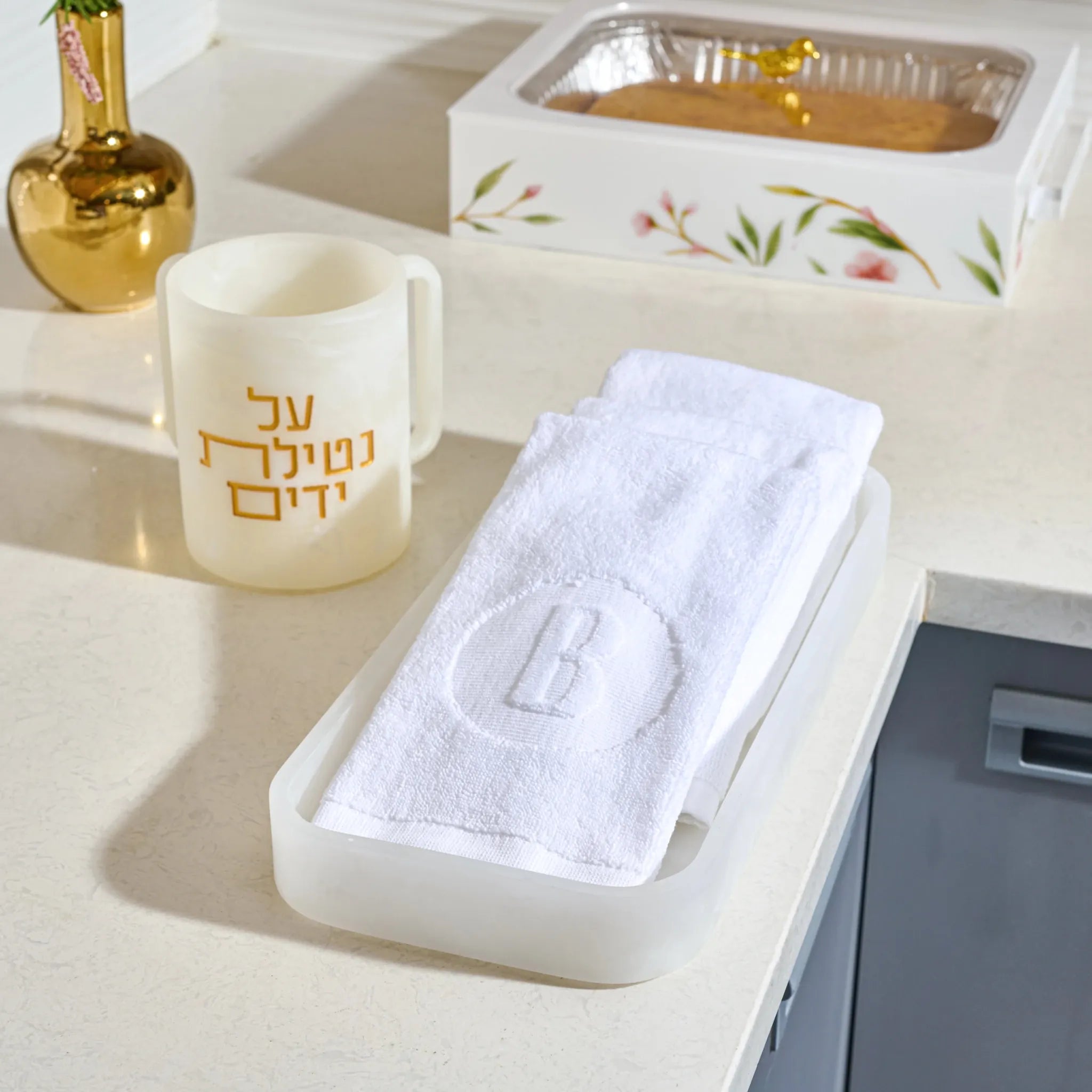A white folded towel with an embroidered letter 'B' sits in a translucent resin holder on a light beige countertop. A frosted resin cream cup with gold Hebrew text is nearby, with a decorative box in the background.