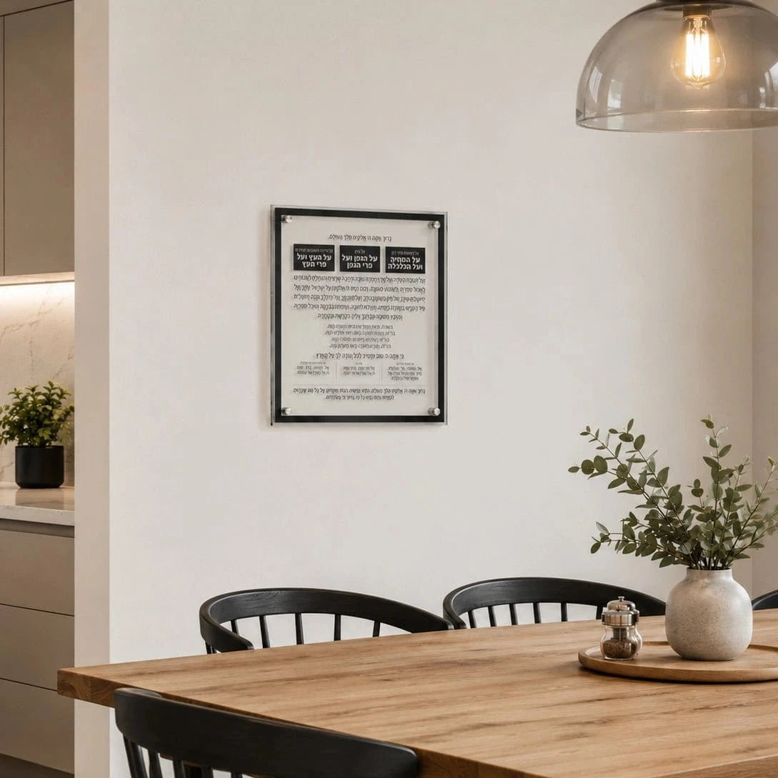 A framed Hebrew and English prayer plaque with a black border hangs on a white wall in a modern dining room setting with a wooden table, black chairs, and a hanging glass dome lamp.