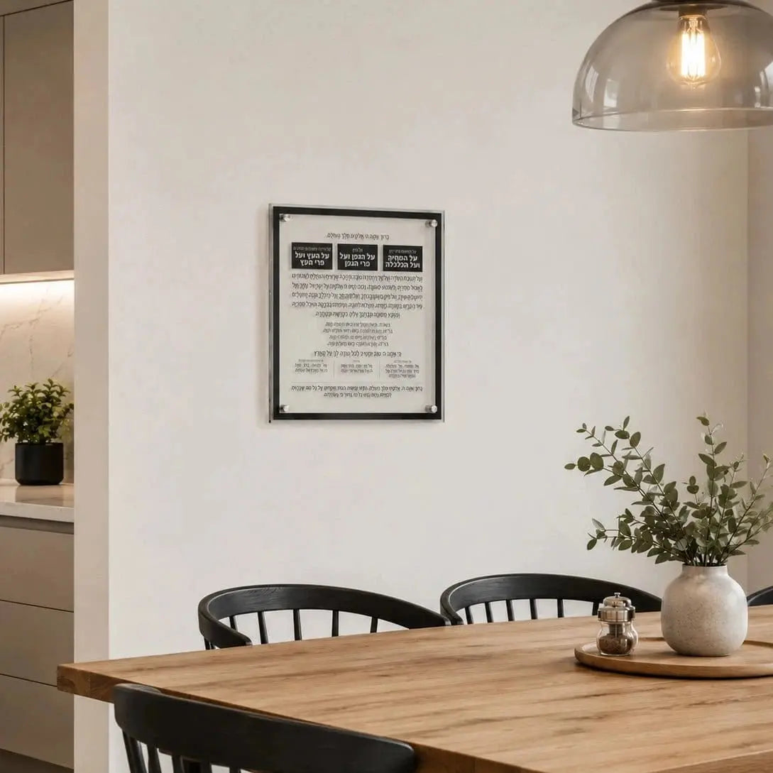 A framed Hebrew and English prayer plaque with a black border hangs on a white wall in a modern dining room setting with a wooden table, black chairs, and a hanging glass dome lamp.