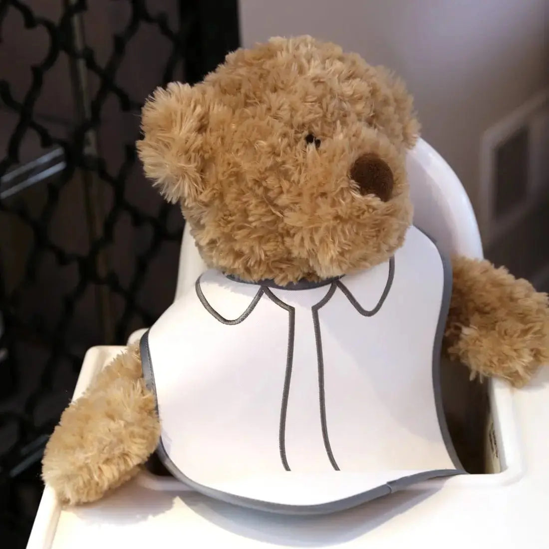 Tan teddy bear wearing a white crocodile patent leather bib with embroidered details, displayed against a neutral background.