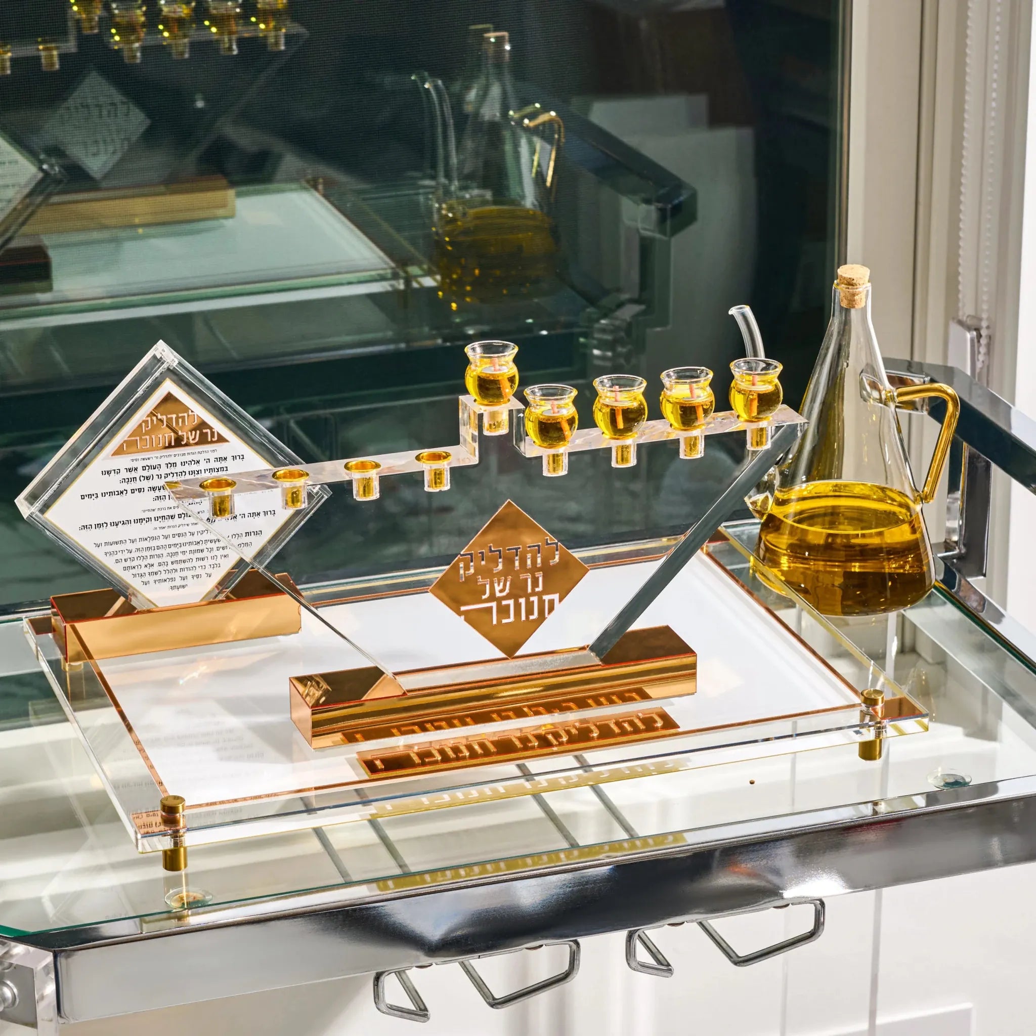 An acrylic and gold Hanukkah menorah set on a glass shelf, featuring a menorah with small clear cups filled with oil, a clear oil bottle, a diamond-shaped plaque with Hebrew and English text, and a central gold sign.