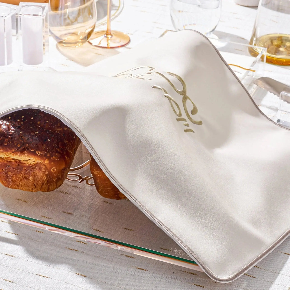 Silver leather Challah cover with gold accents displayed on a white table with bread and wine, perfect for Shabbat.