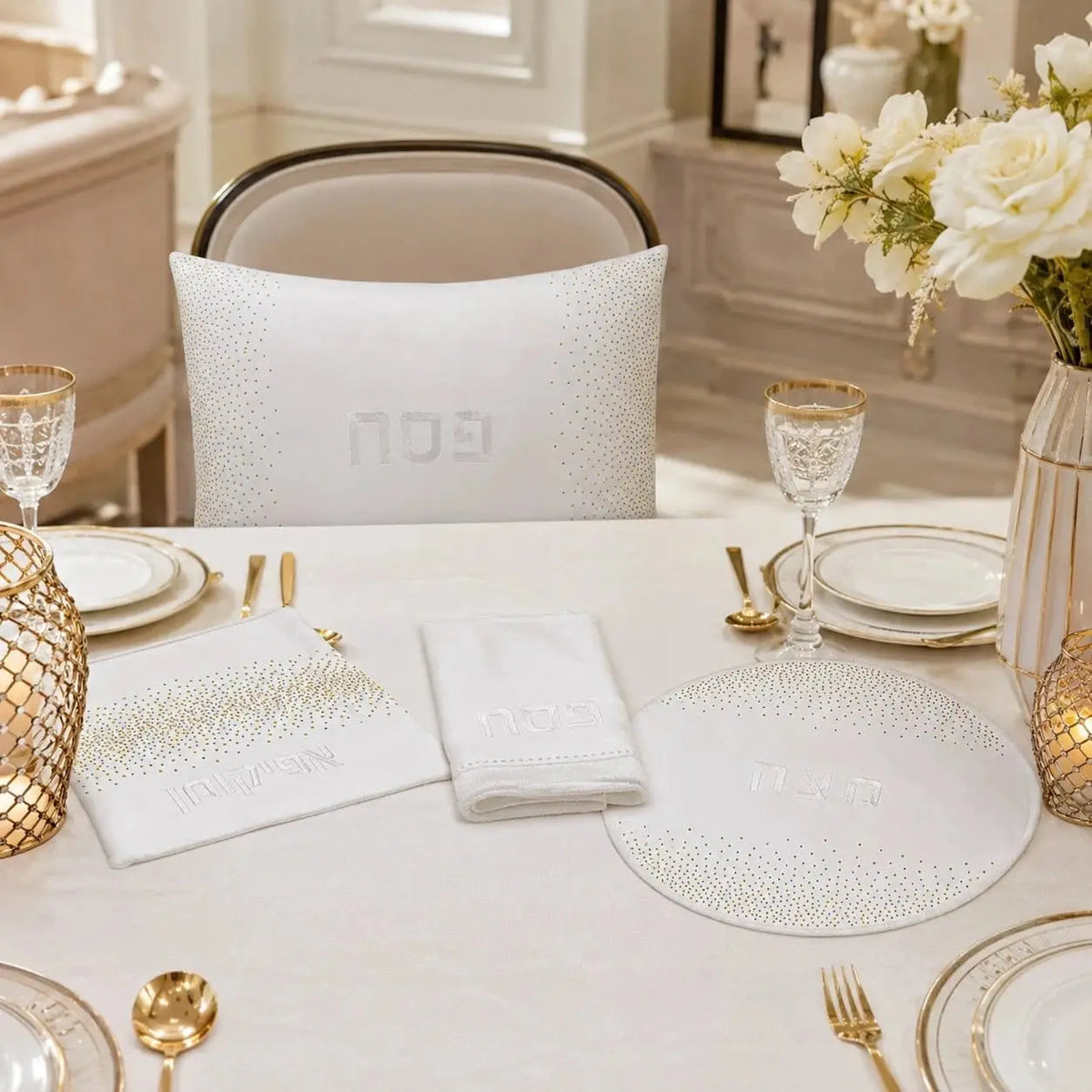 A styled dining table setting featuring a white embroidered pesah set cushion, matching white napkins, a white placemat, and gold-accented tableware in a bright, elegant room.