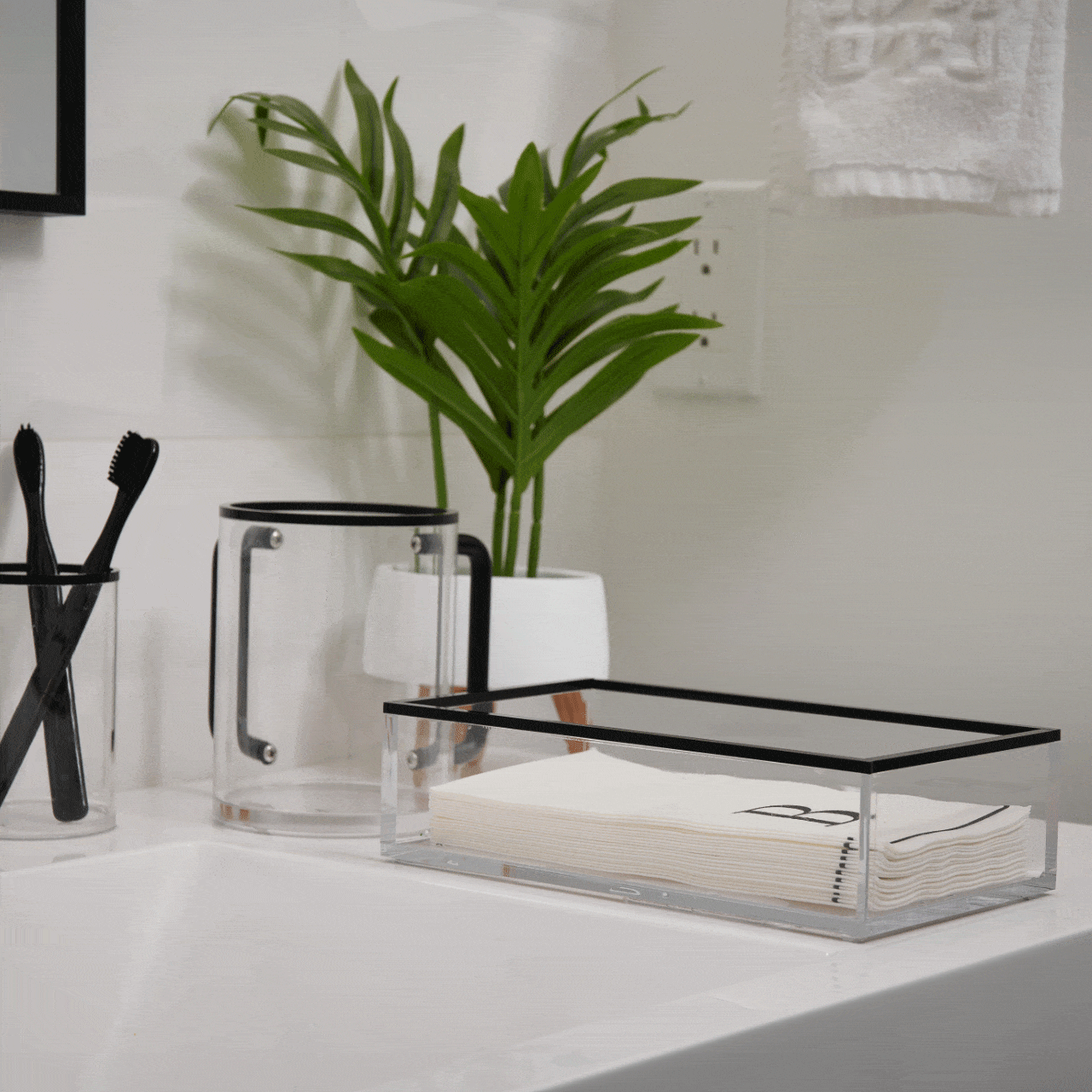 Clear acrylic Edge Guest Towel Holder with black trim displayed on white desk next to plant, brush holder, and notepad in modern bathroom setting.