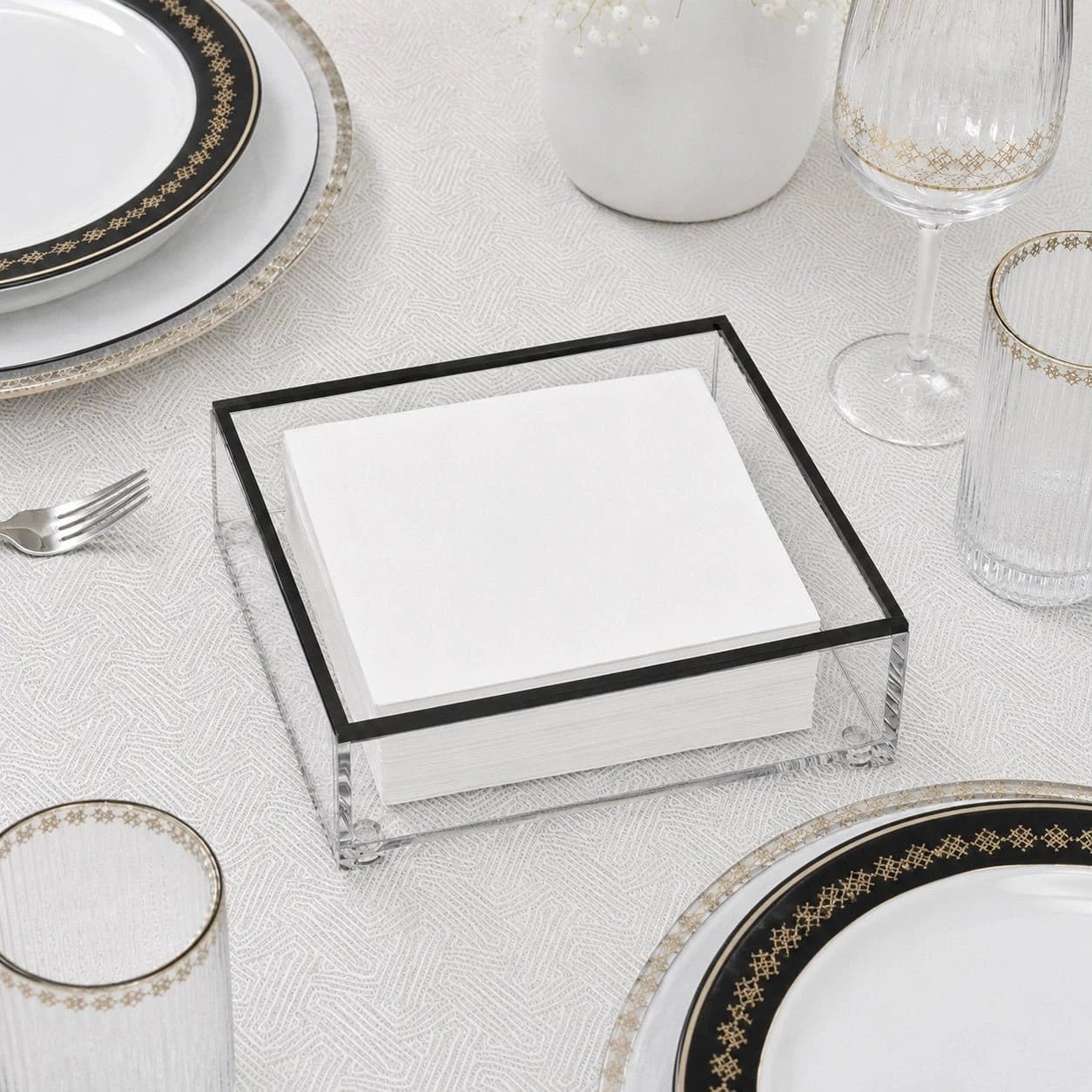 A clear square napkin holder with a black frame, filled with white folded napkins, styled on a white tablecloth with gold-rimmed plates, glasses, and a fork.
