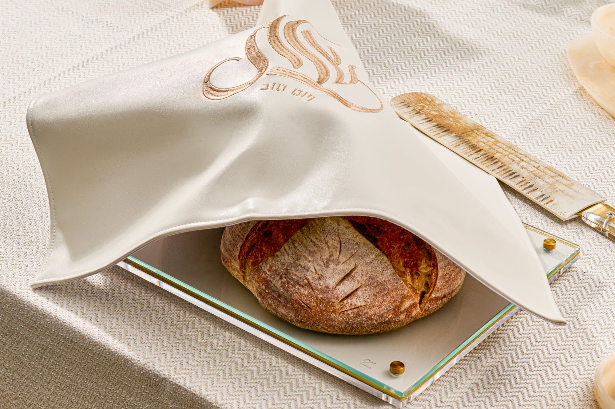 Embroidered regal challah board with an intricate gold and white floral pattern under a protective acrylic surface on a white marble table.