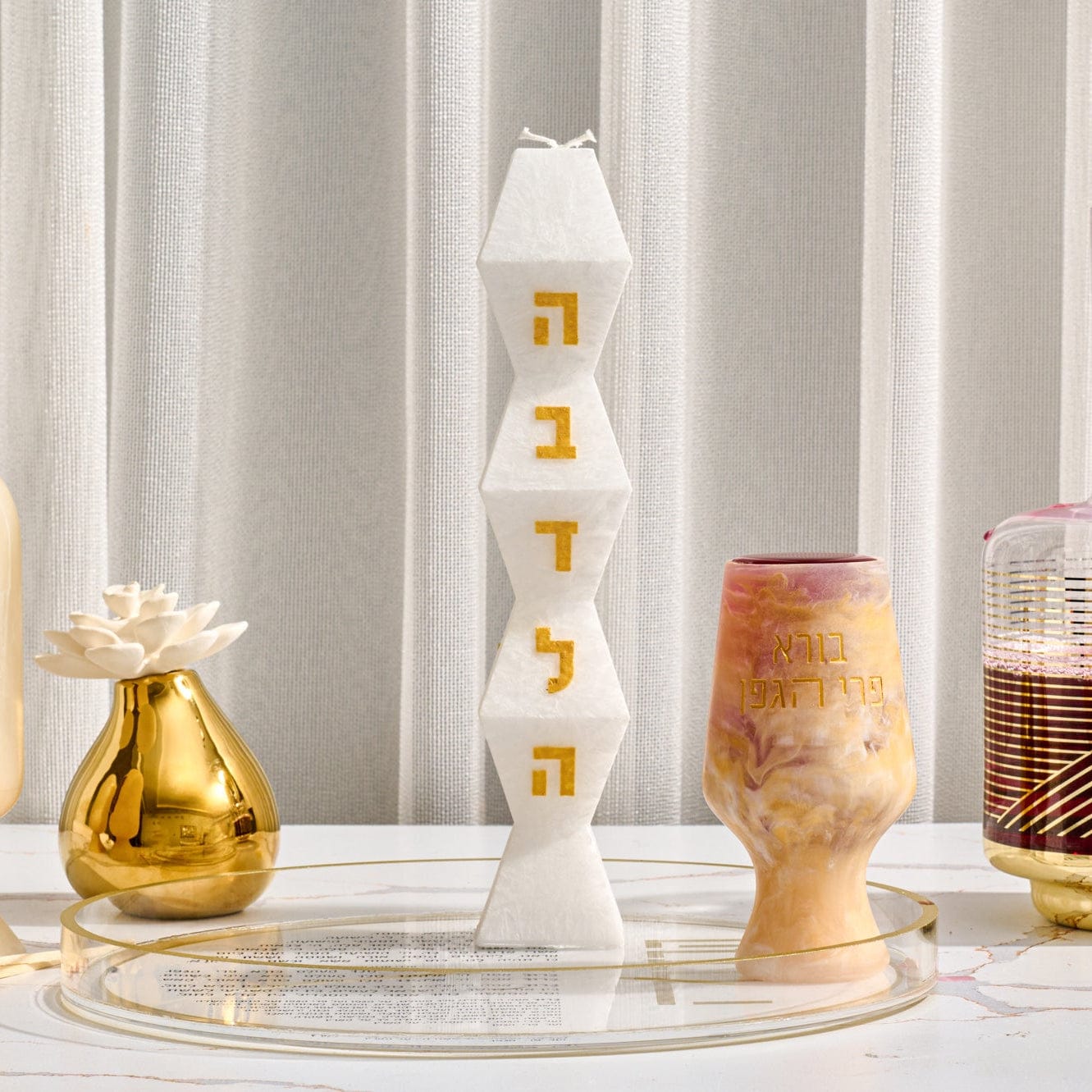 Facet Havdalah candle in matte white with geometric stacked design and gold Hebrew lettering, displayed on clear round tray alongside decorative Judaica pieces in an elegant modern setting.