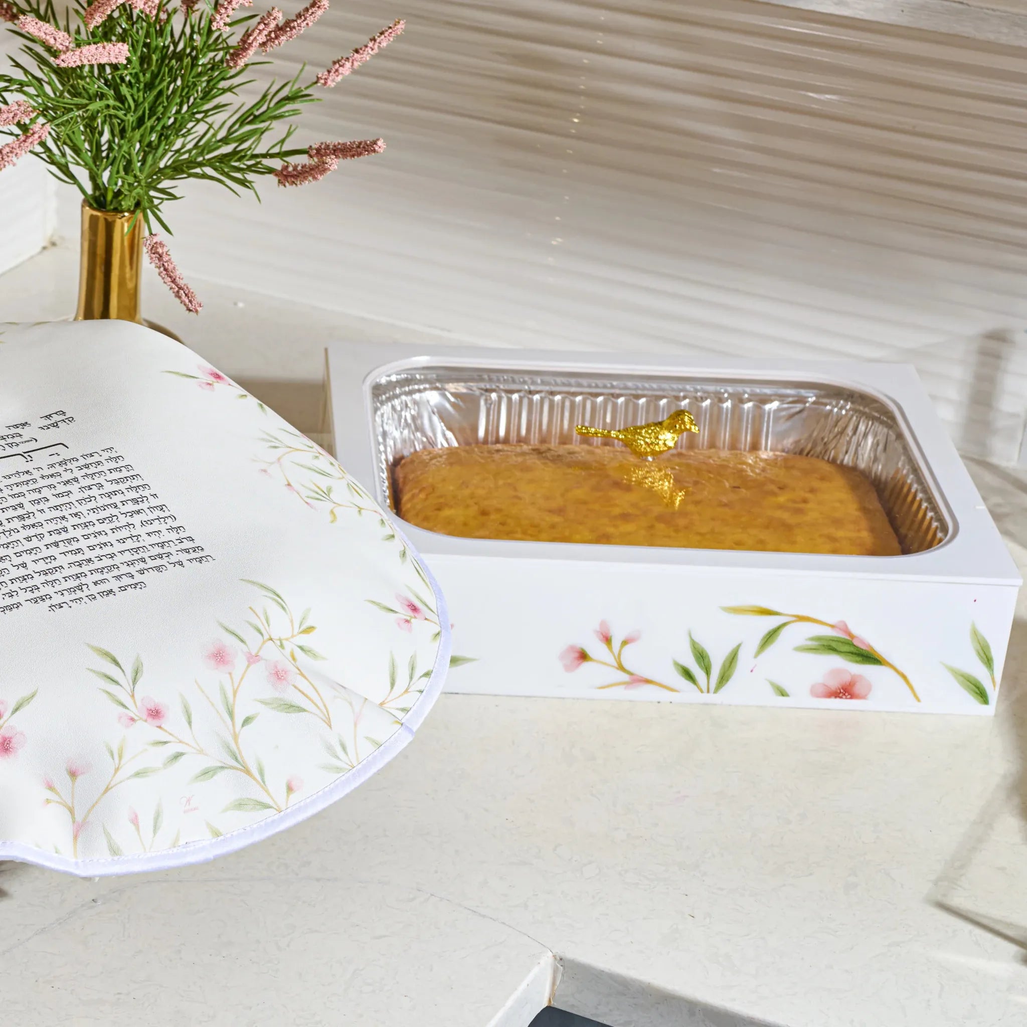 light countertop, with a rectangular foil pan in a white acrylic floralpan holder holding a golden bird with a cake inside