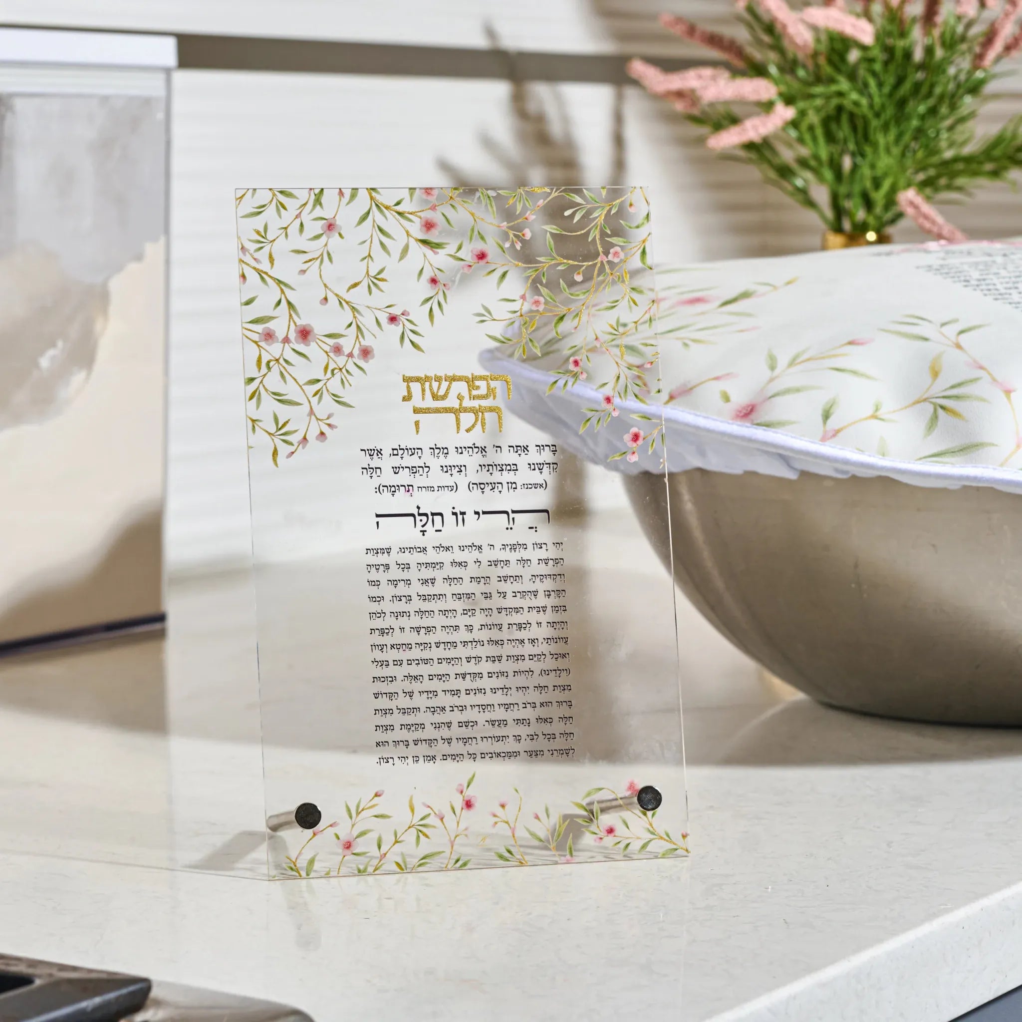 A decorative printed card with floral and leaf patterns in soft green, pink, and gold, featuring Hebrew text at the top and a block of smaller text below, displayed on a clear stand in a kitchen setting.
