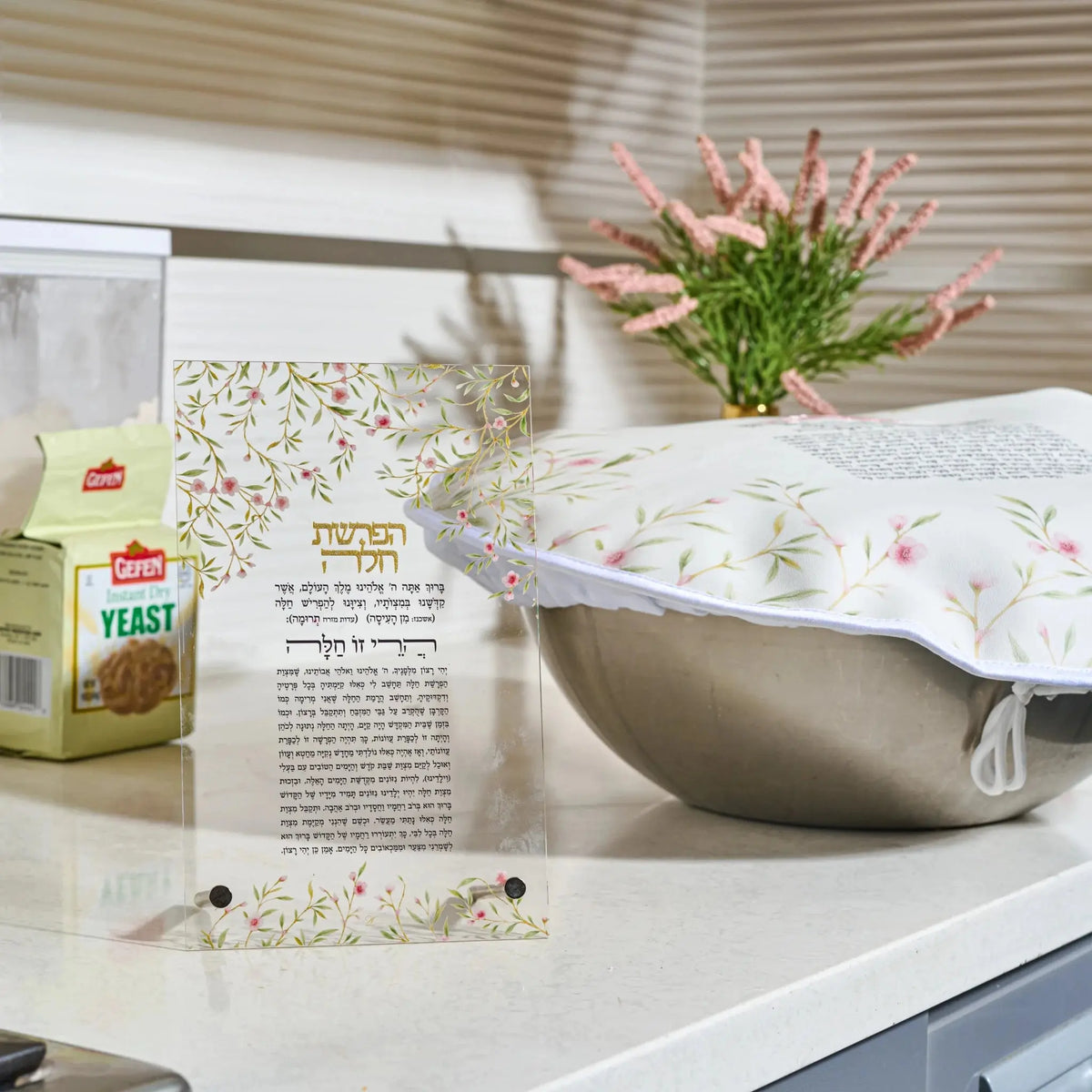 A decorative Challah dough cover over with Hebrew text and a floral border stands on a kitchen counter next to a box of Leavened yeast. In the background, a white electric blanket with a matching floral pattern is visible, along with a vase of pink flowers and window blinds.