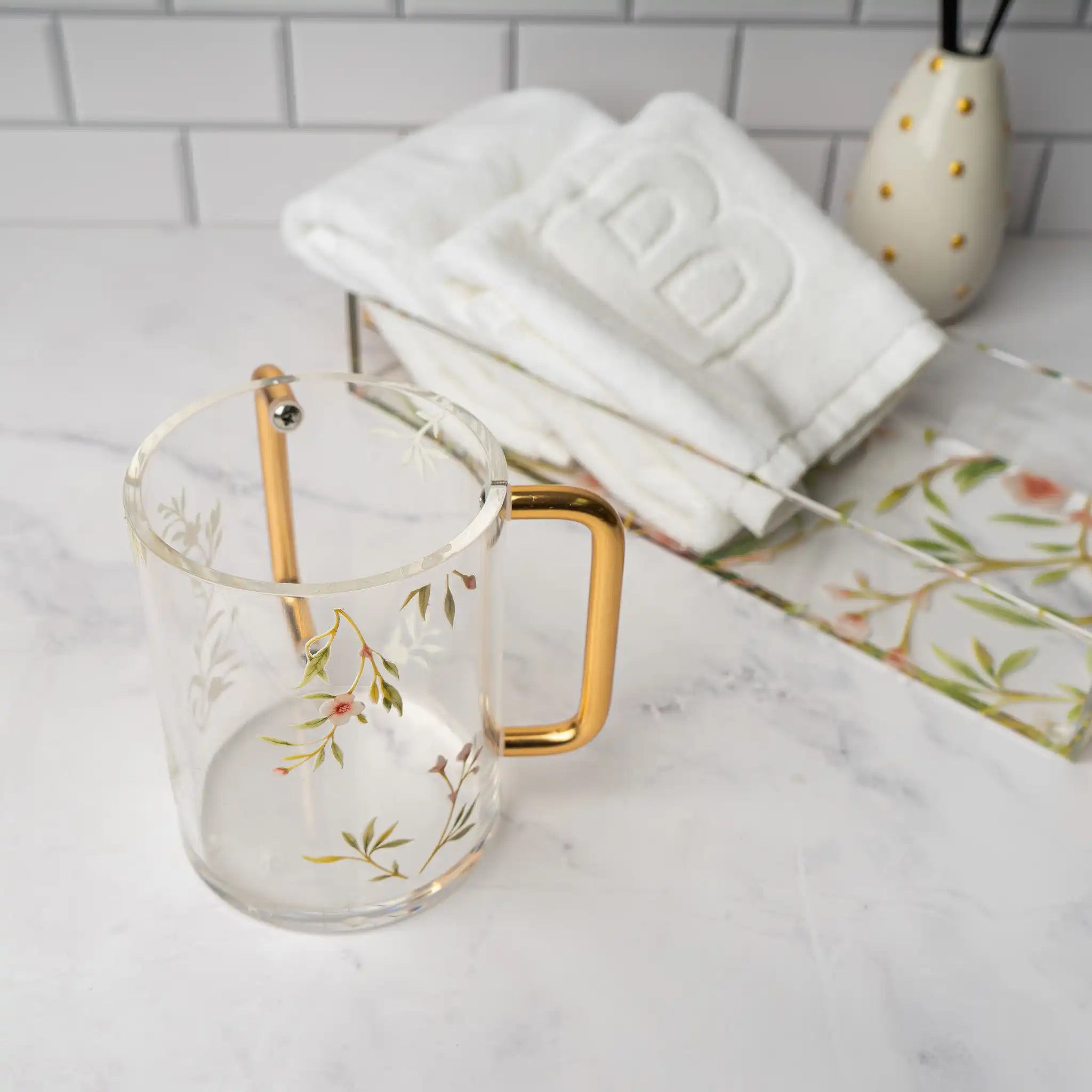 Lucite Washing cup with pink floral design and rose gold handle on a marble countertop with towels in the back - Waterdale Collection