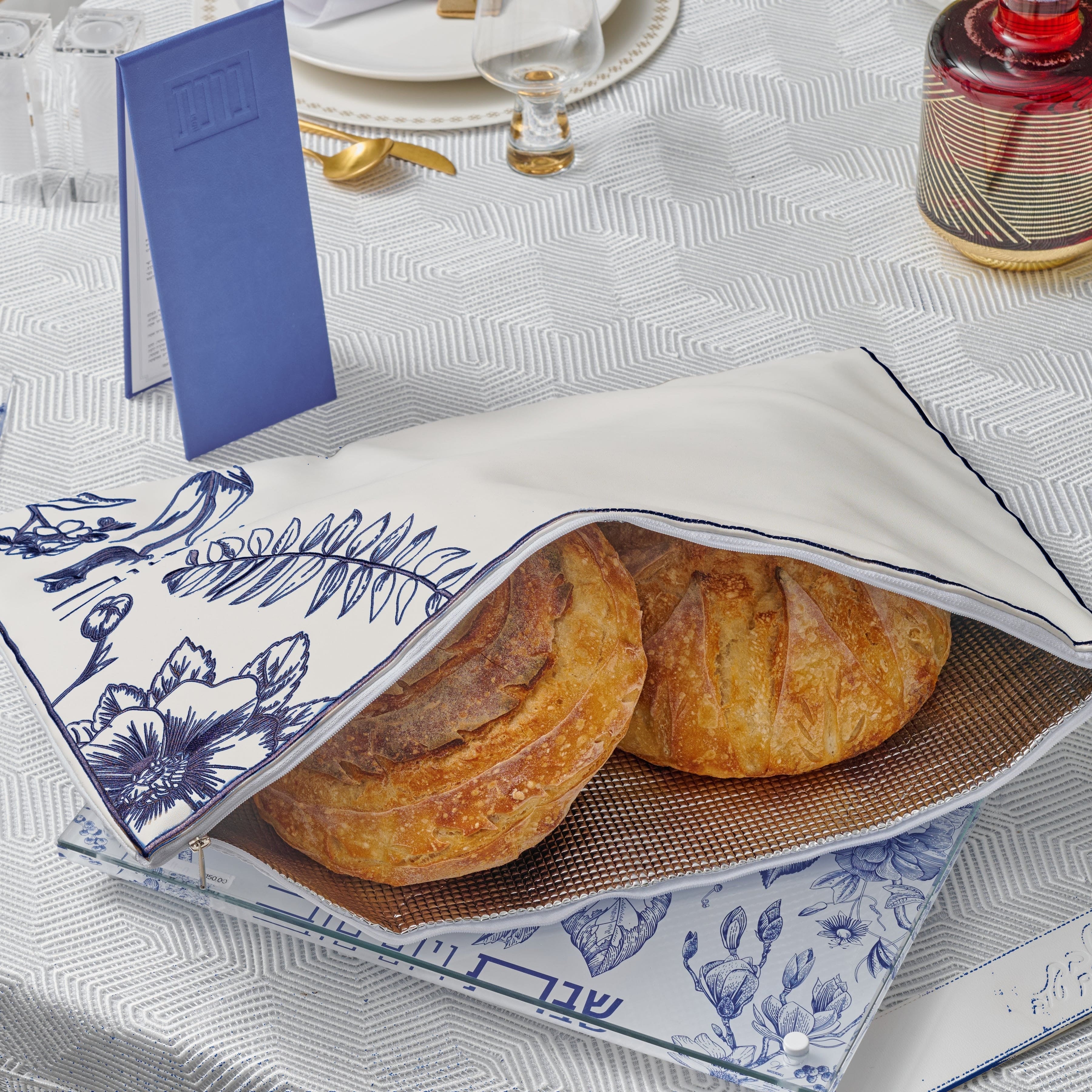 Blue French Toile Challah Pouch Cover styled on an elegant Shabbat table with gold-accented tableware and a blue bencher booklet.