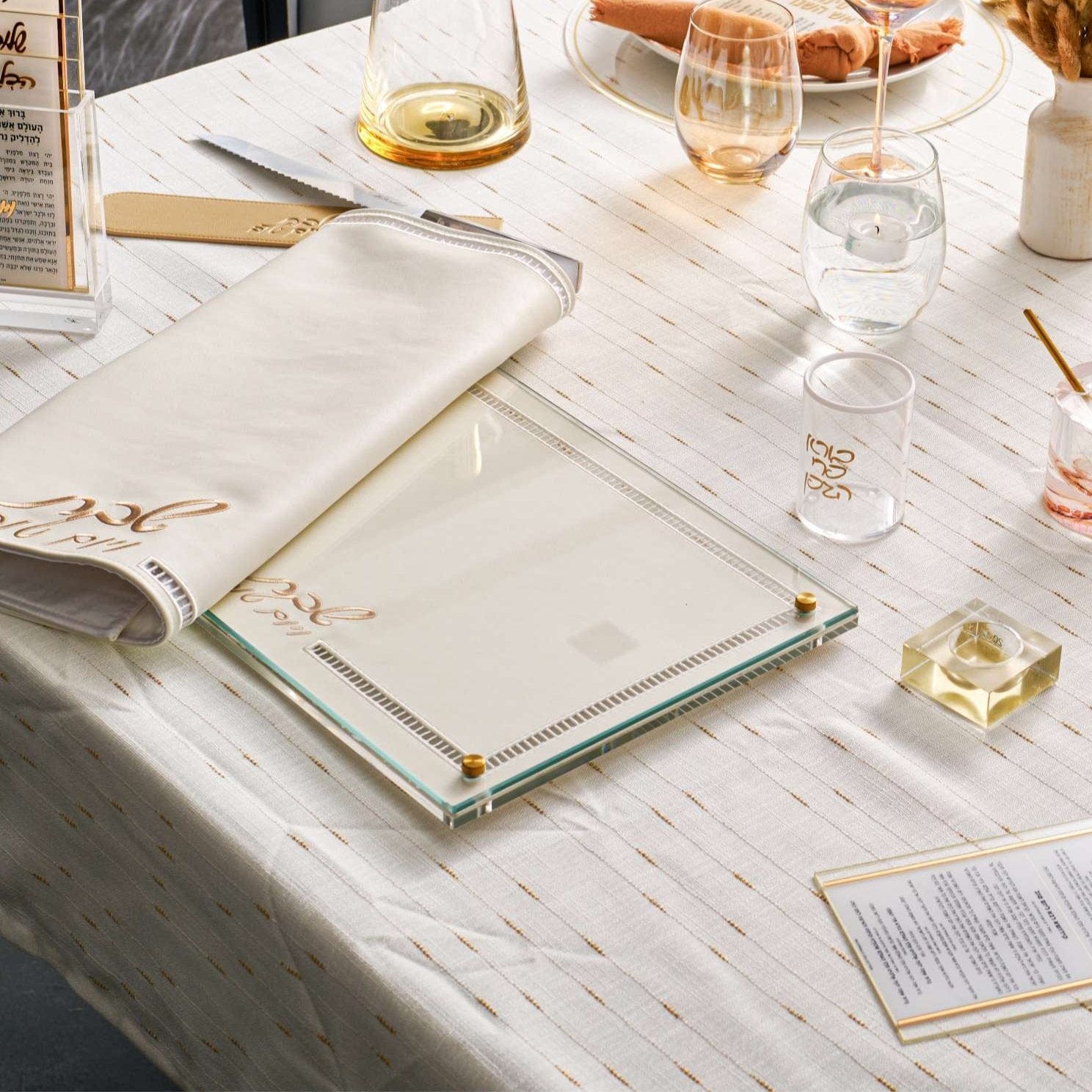 Elegant Shabbat table setting with a Hemstich Challah Cover and a glass challah board on a white and gold tablecloth.