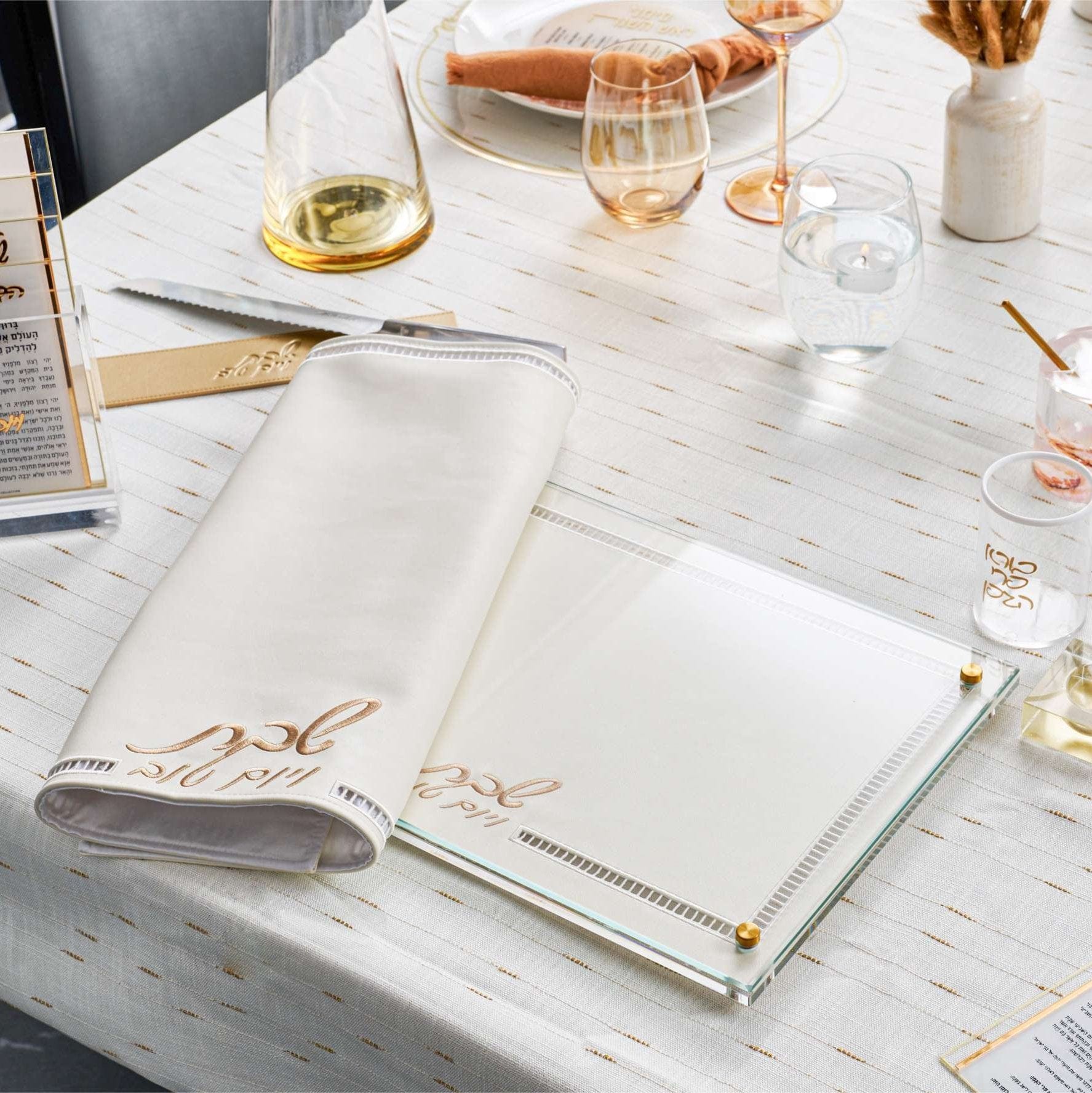 Elegant Shabbat table setting with a Hemstich Challah Cover and a glass challah board on a white and gold tablecloth.