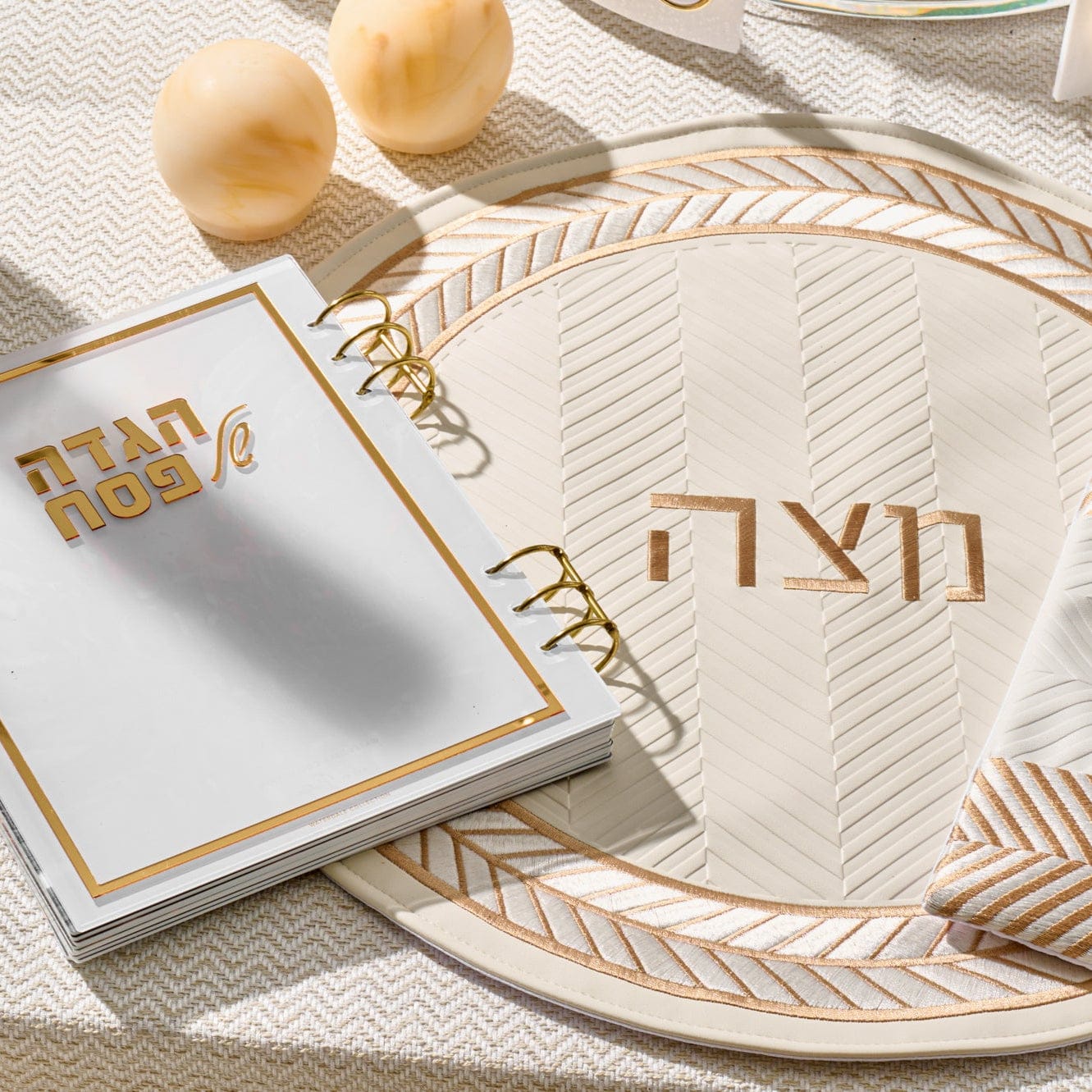 Herringbone matzah cover in cream with gold accents and Hebrew “Matzah” lettering, displayed on a coordinating Seder plate.