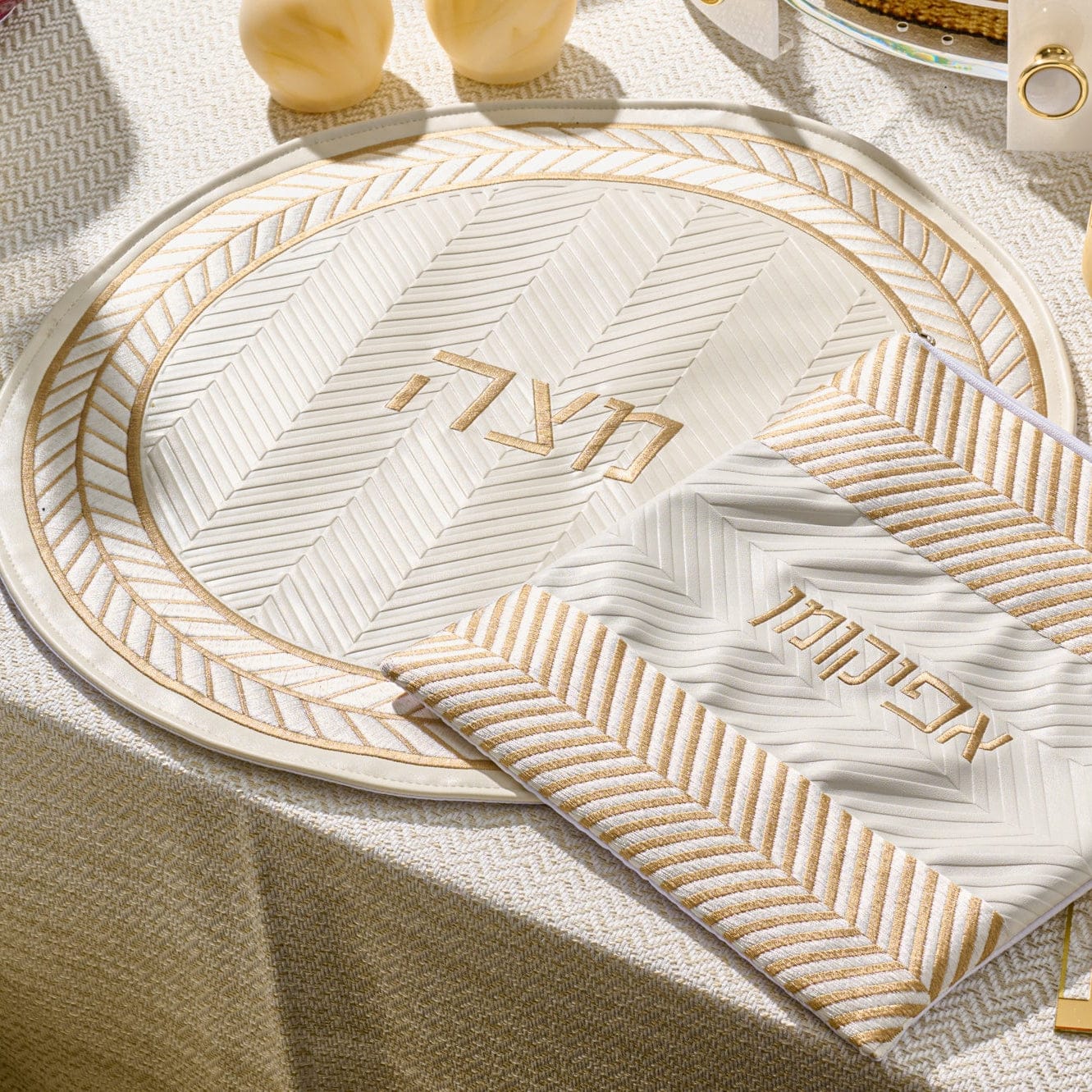 Herringbone matzah cover in cream with gold accents and Hebrew “Matzah” lettering, displayed on a matching Seder plate on a Pesach table.