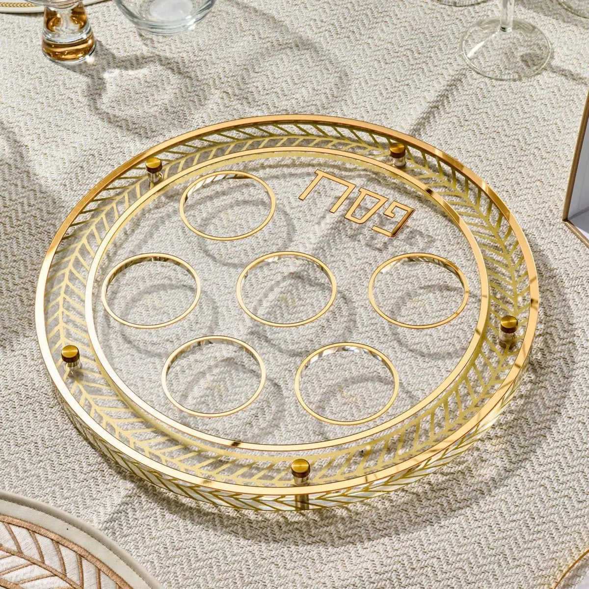 Herringbone Seder plate with clear lucite wells and gold-tone metal rim, displayed on a patterned tablecloth for Passover.