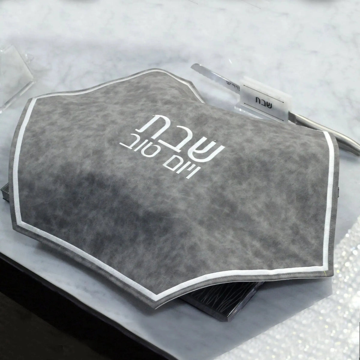 Marbleized gray hexagon challah cover in leather with embroidered Hebrew text, displayed on white surface.
