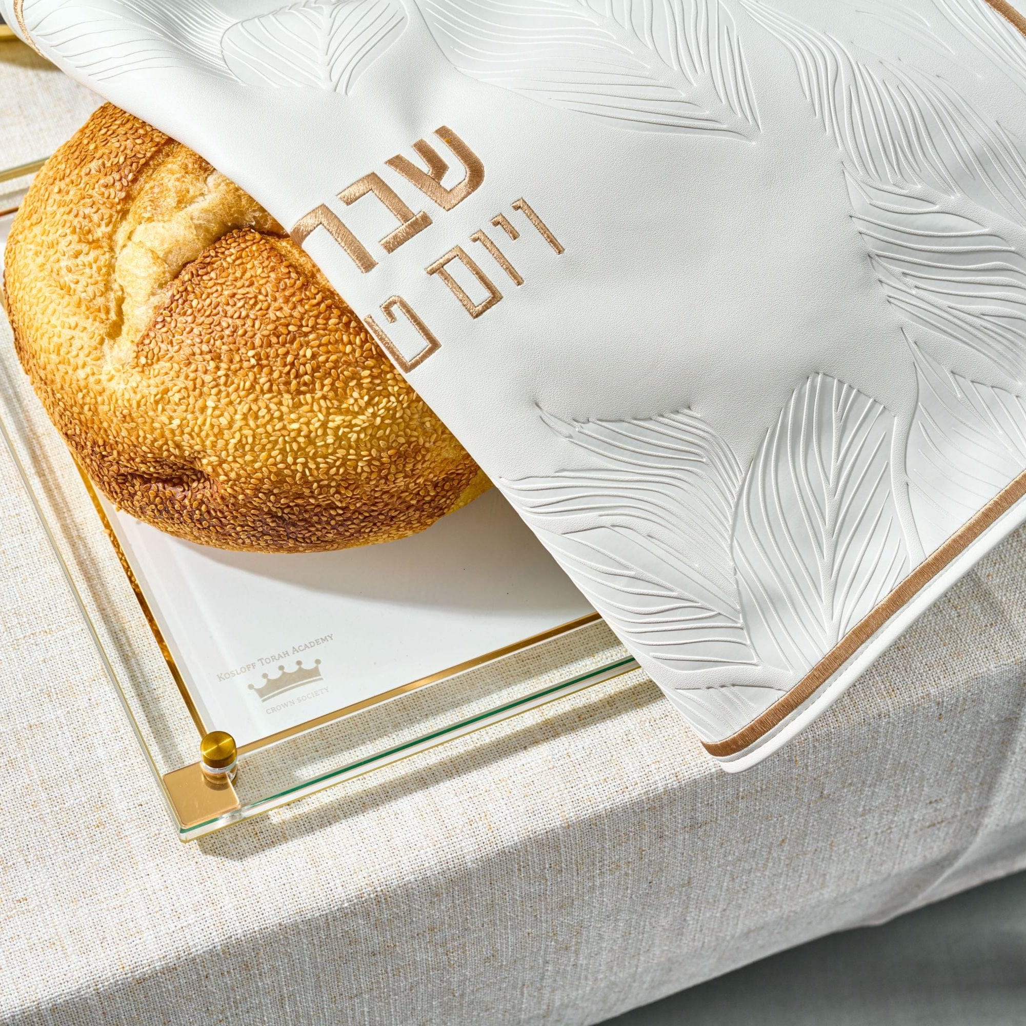 Round challah bread partially covered with Leaf Challah Cover on a glass challah board.