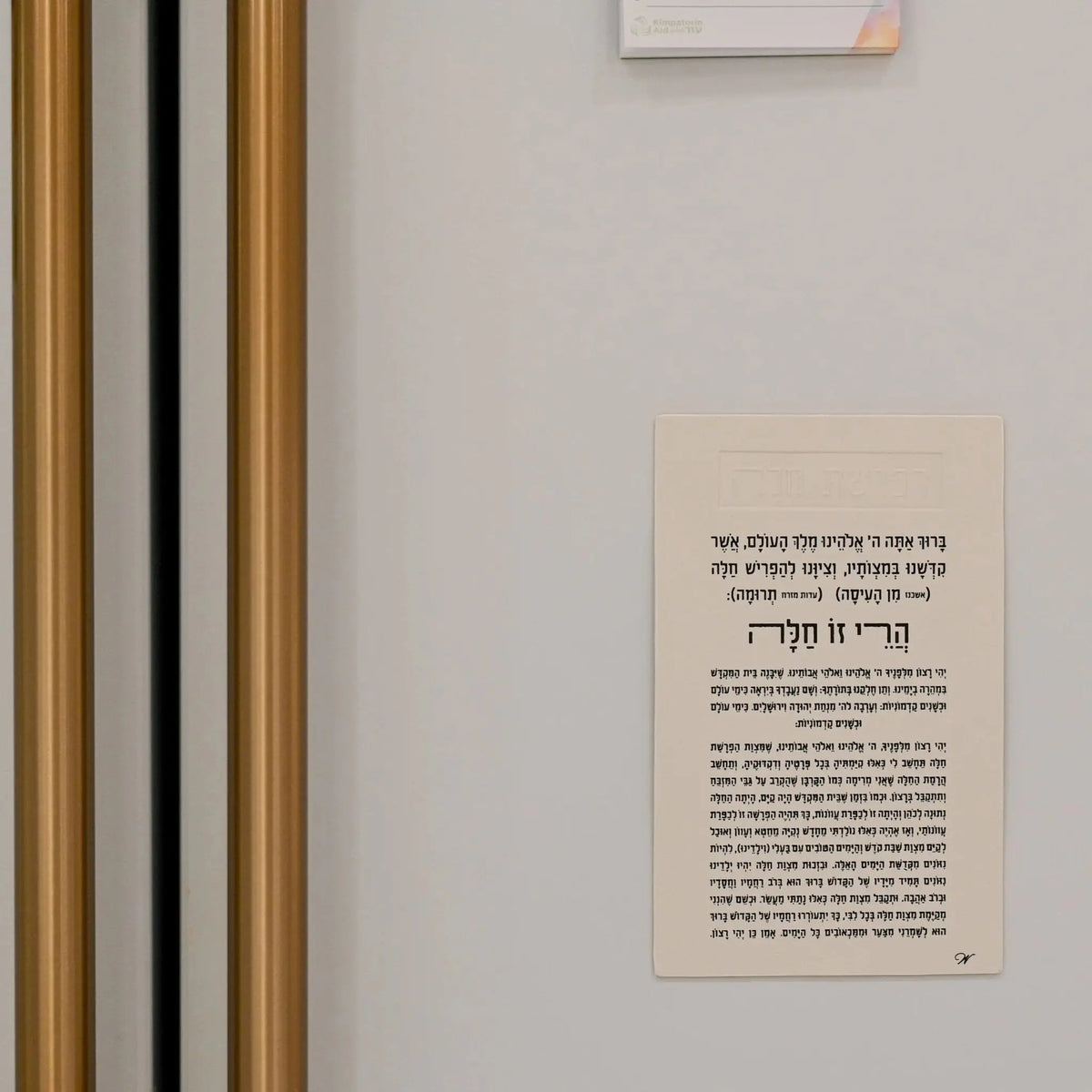 Leather Hafrashas Challah magnet in black leatherette with Hebrew text for Shabbat blessings, displayed on white wall near framed striped artwork.