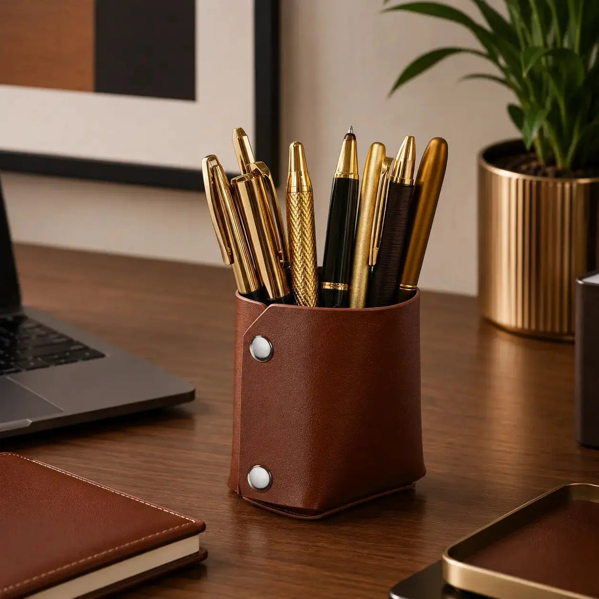 Brown leather pen cup with gold pens displayed on a wooden desk next to a laptop and potted plant, perfect for office organization.