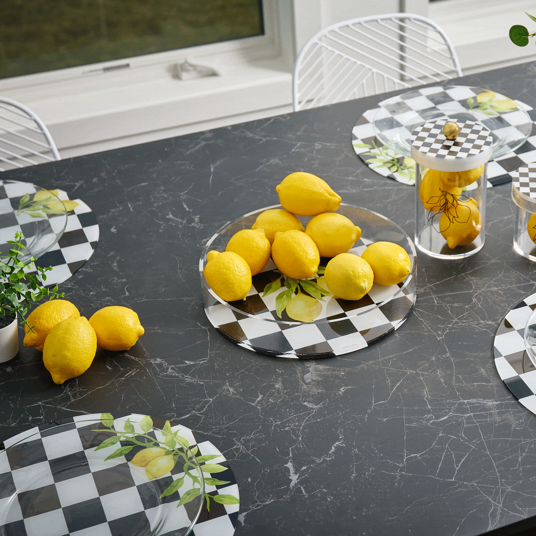Lemona chargers with black and white checkered pattern featuring lemon motifs, displayed on dark marble table with fresh lemons and glassware.