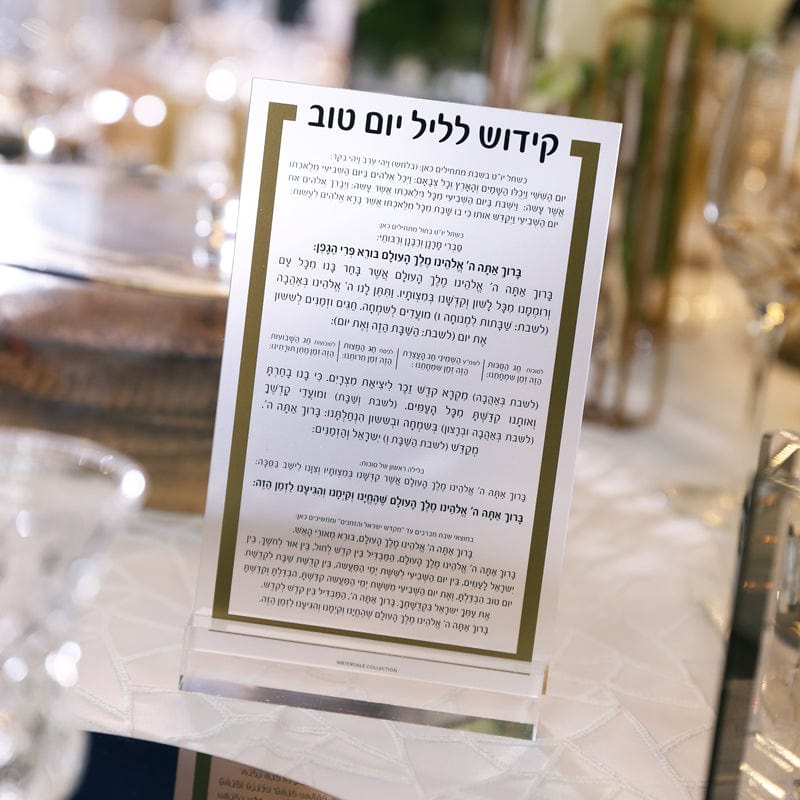 A clear lucite Yom Tov Kiddush card by Waterdale Collection, standing on a white textured tablecloth next to luxury dinnerware and gold-trimmed decor.