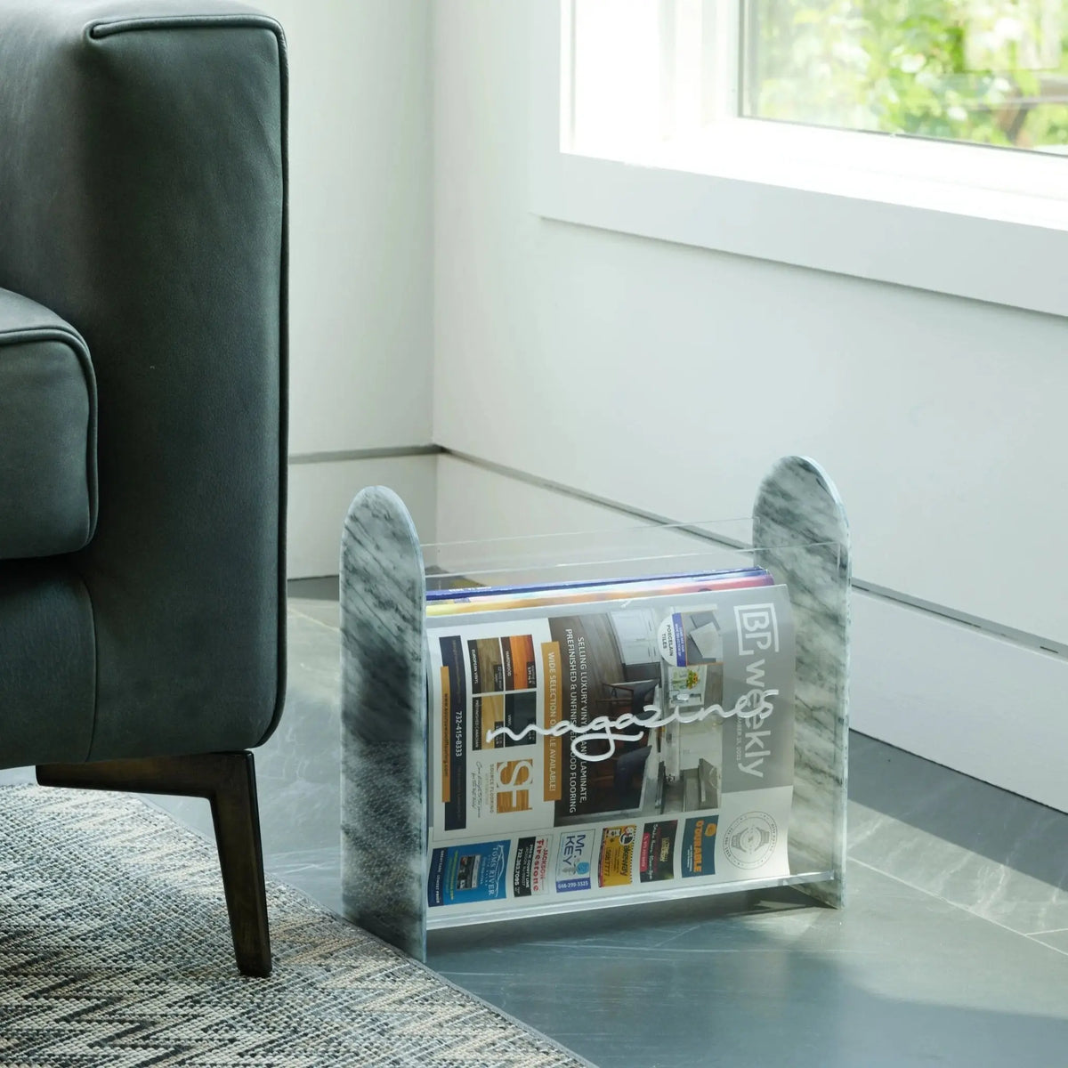 Clear acrylic magazine rack holding publications and decorative items, displayed in a modern living room setting near a black chair.