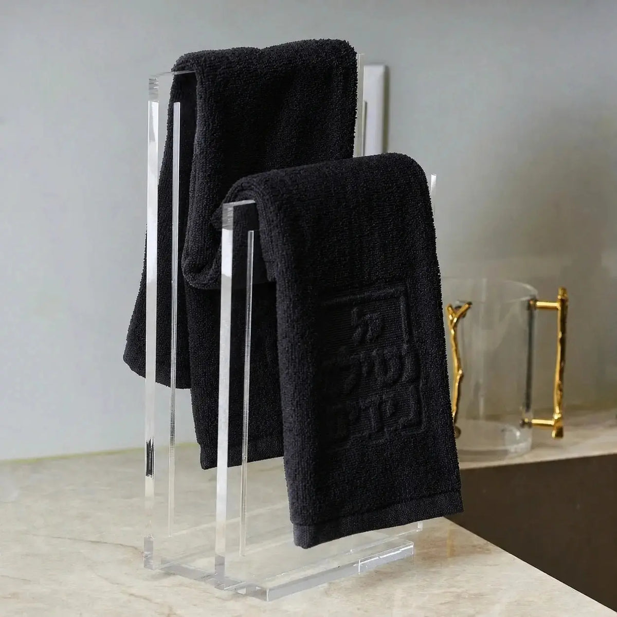 Embossed netilas yadayim towel set displayed on modern metal stand with three folded towels and decorative gold item on shelf.