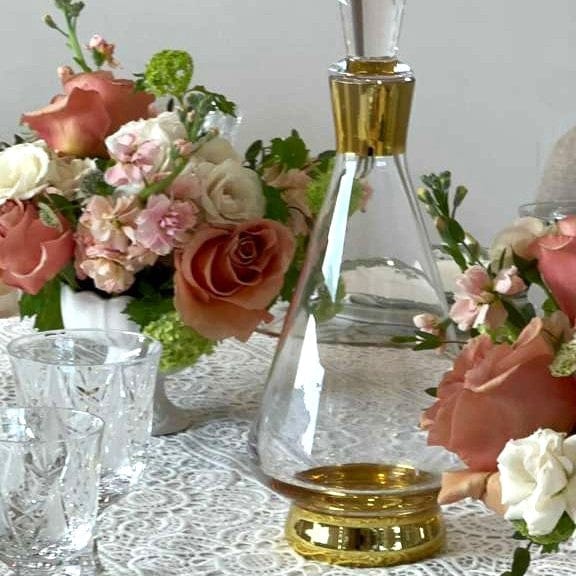 Modern glass wine decanter with gold-rimmed accents and a tapered neck, displayed on a lace tablecloth next to elegant floral arrangements and crystal glasses.
