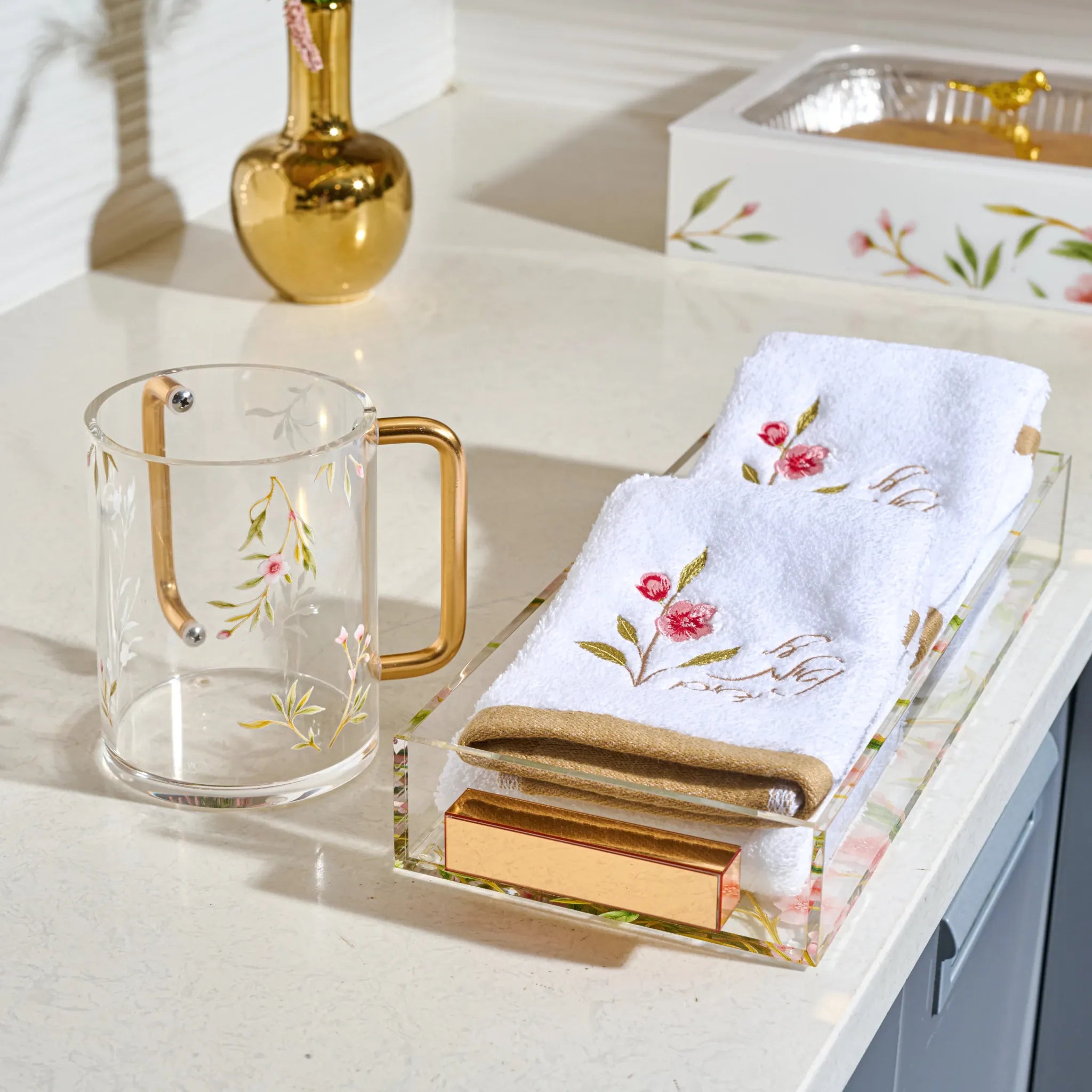 A styled tabletop scene with a clear acrylic tray holding folded white embroidered towels and a gold-toned soap bar, alongside a clear glass mug with a gold handle and floral print. A gold vase and a floral-patterned box are also visible in the background on a light countertop.