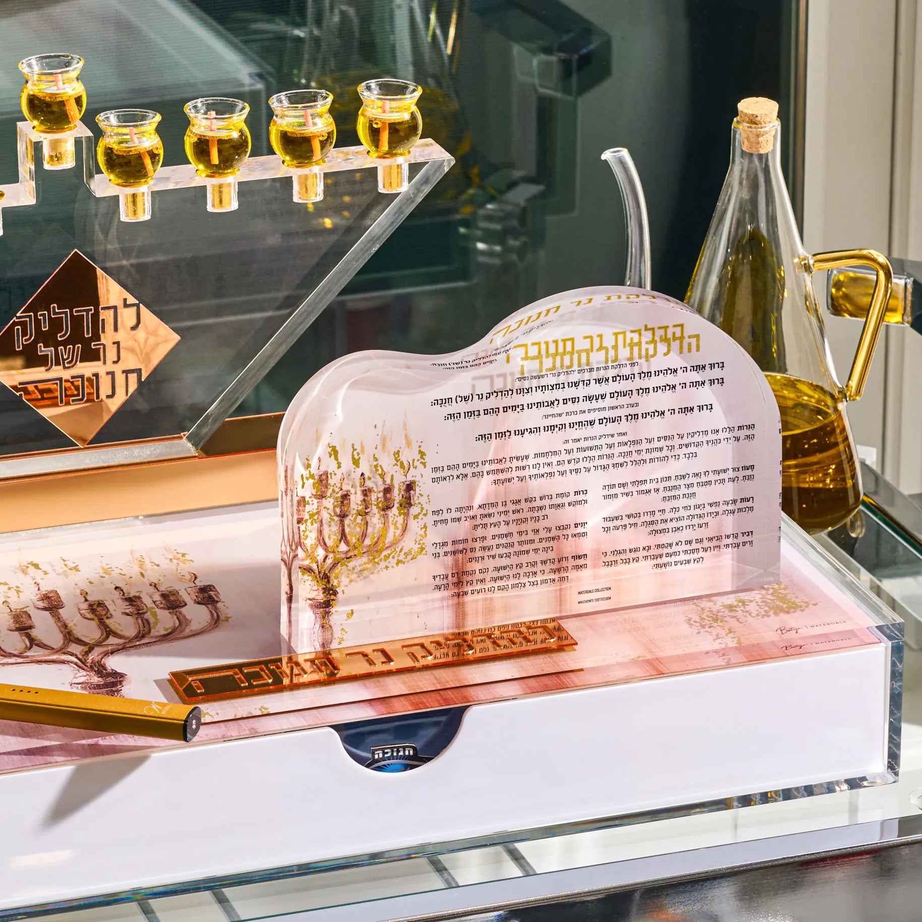 A Hanukkah menorah set displayed on a glass shelf, featuring a clear acrylic menorah with a printed menorah illustration and Hebrew text, alongside a gold-colored metal menorah, a gold candlestick, a glass bottle of olive oil, and small glass cups.