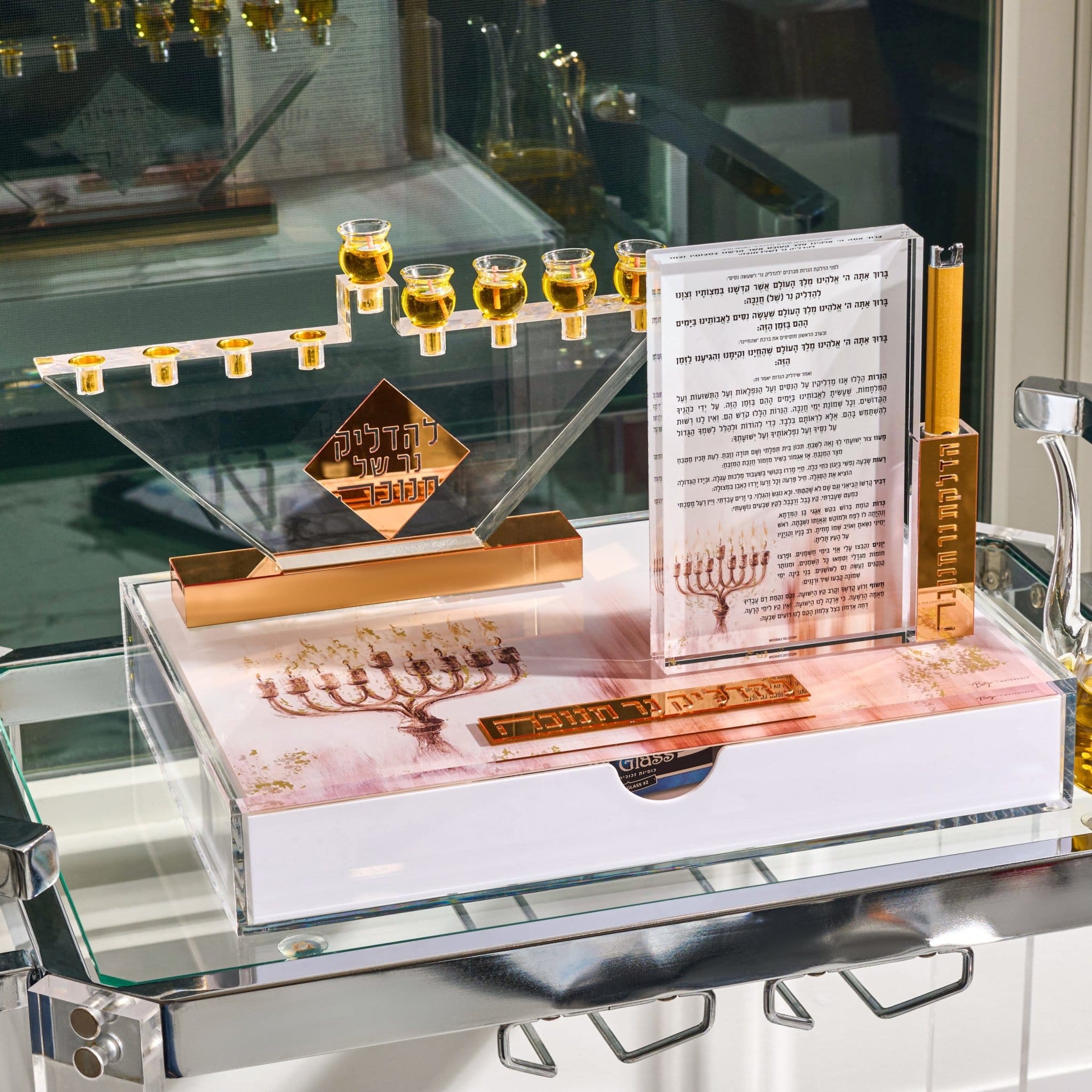 A decorative Hanukkah menorah set displayed on a glass shelf, featuring a clear acrylic menorah with gold-toned candle holders, a matching gold-toned base, a printed instruction card in Hebrew and English, a gold-toned candle, and a white box with a menorah illustration and a gold-toned plaque.