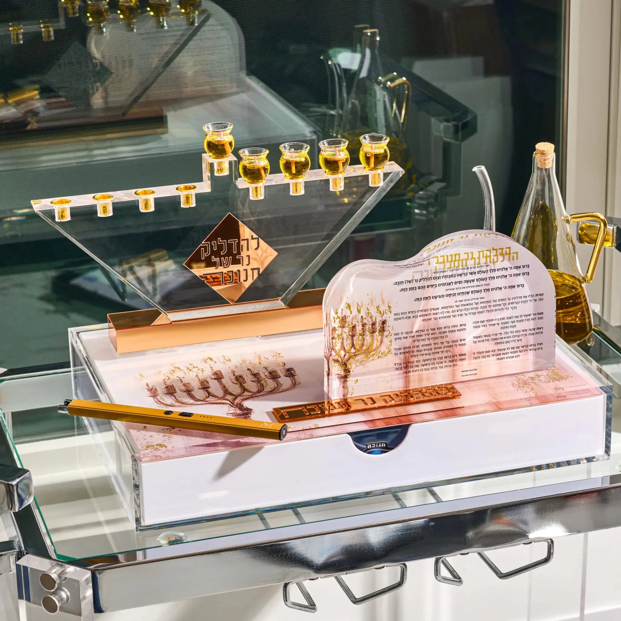 A chanukah candle lighting set displayed on a glass shelf, featuring a clear acrylic menorah with gold-toned candle holders, a gold oil lamp, a white box with a gold lighter , a clear acrylic candle holder with printed Hebrew text, and a gold candlestick.