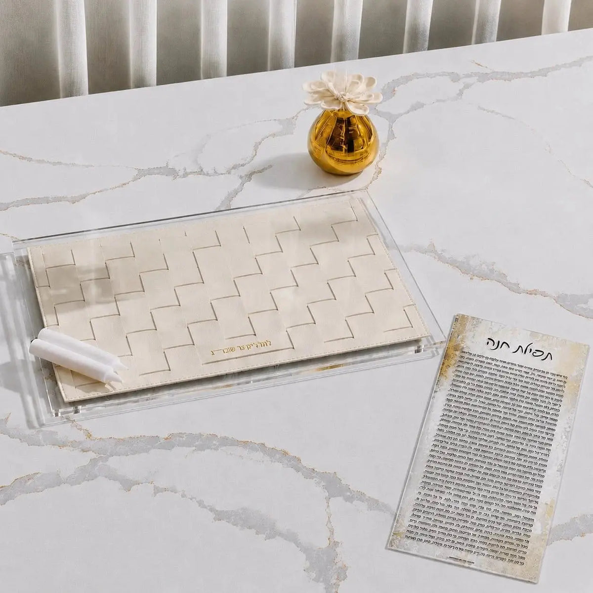 A hadlokas neiros tray set on a white marble table, featuring a clear acrylic base with a beige woven basket insert, two white candles, a small gold decorative vase, and a printed Hebrew tefilas chana card