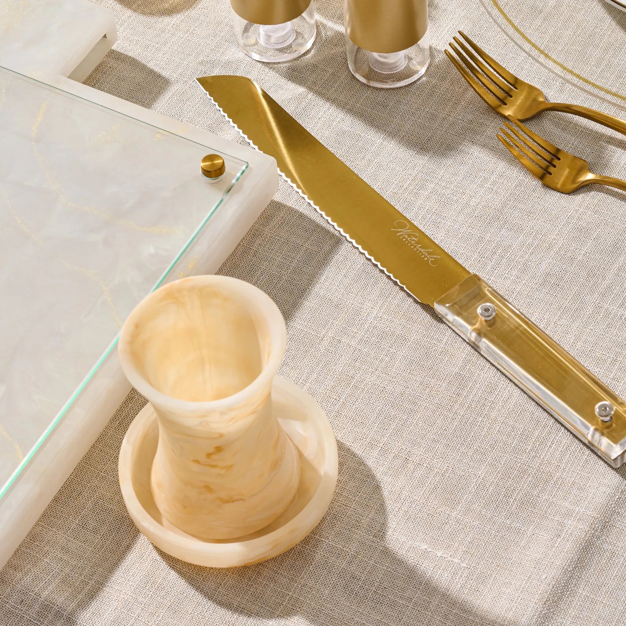 A close-up styled tabletop scene showing a gold-toned knife with a clear handle and a matching gold fork, alongside a gold resin becher